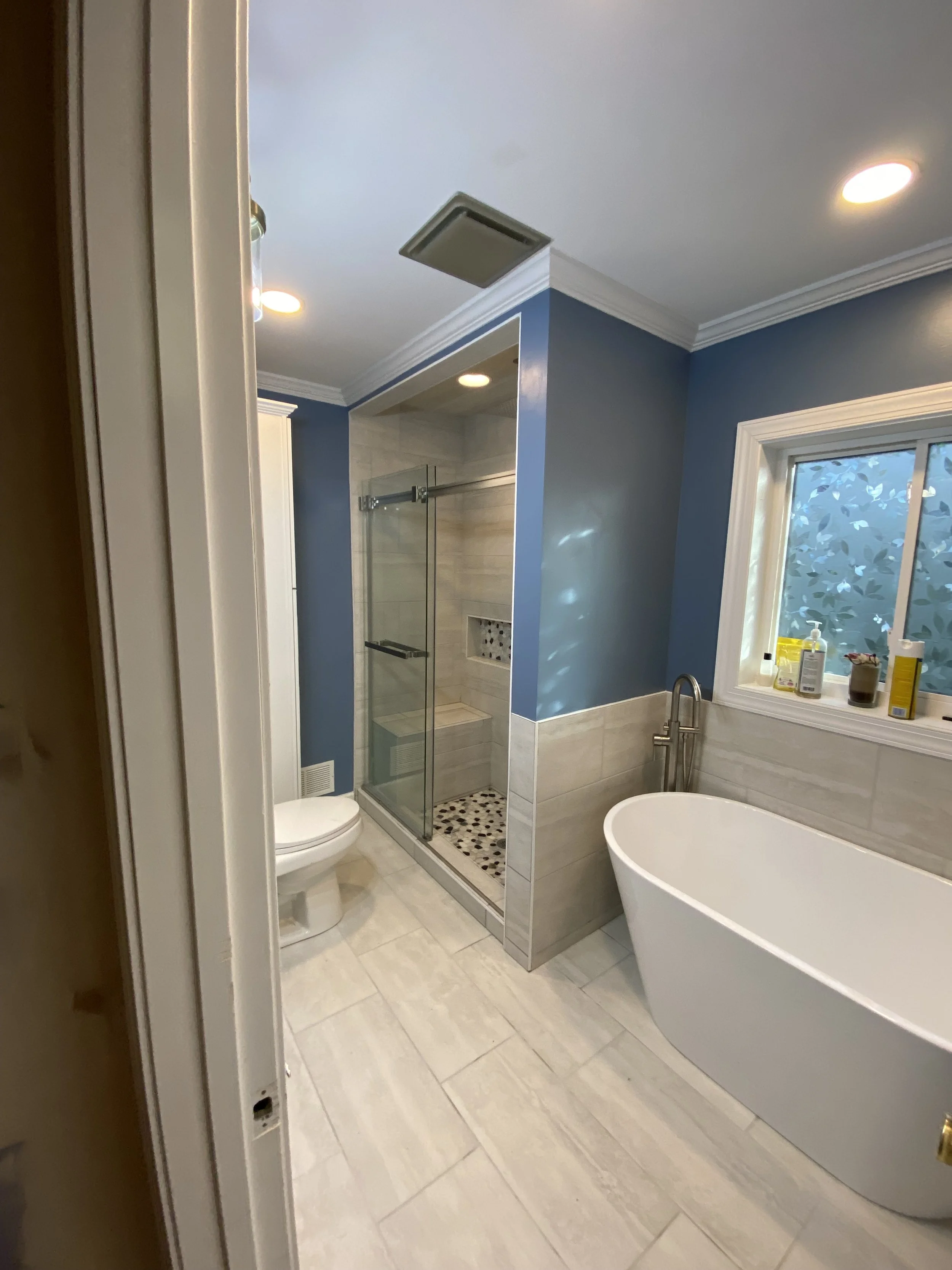 A modern bathroom with a blue accent wall, a freestanding bathtub next to a window, a glass-enclosed shower stall, a toilet, and beige tile flooring and walls.