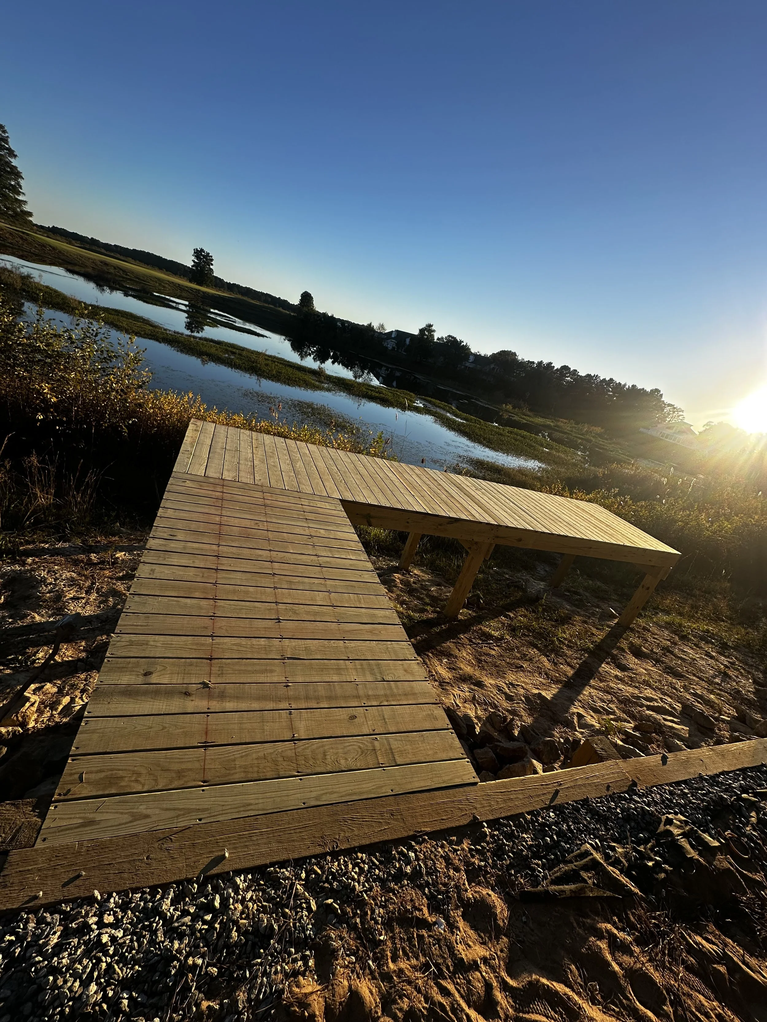 A wooden dock extends over a natural wetland area during sunset, with water, trees, and a clear blue sky in the background.