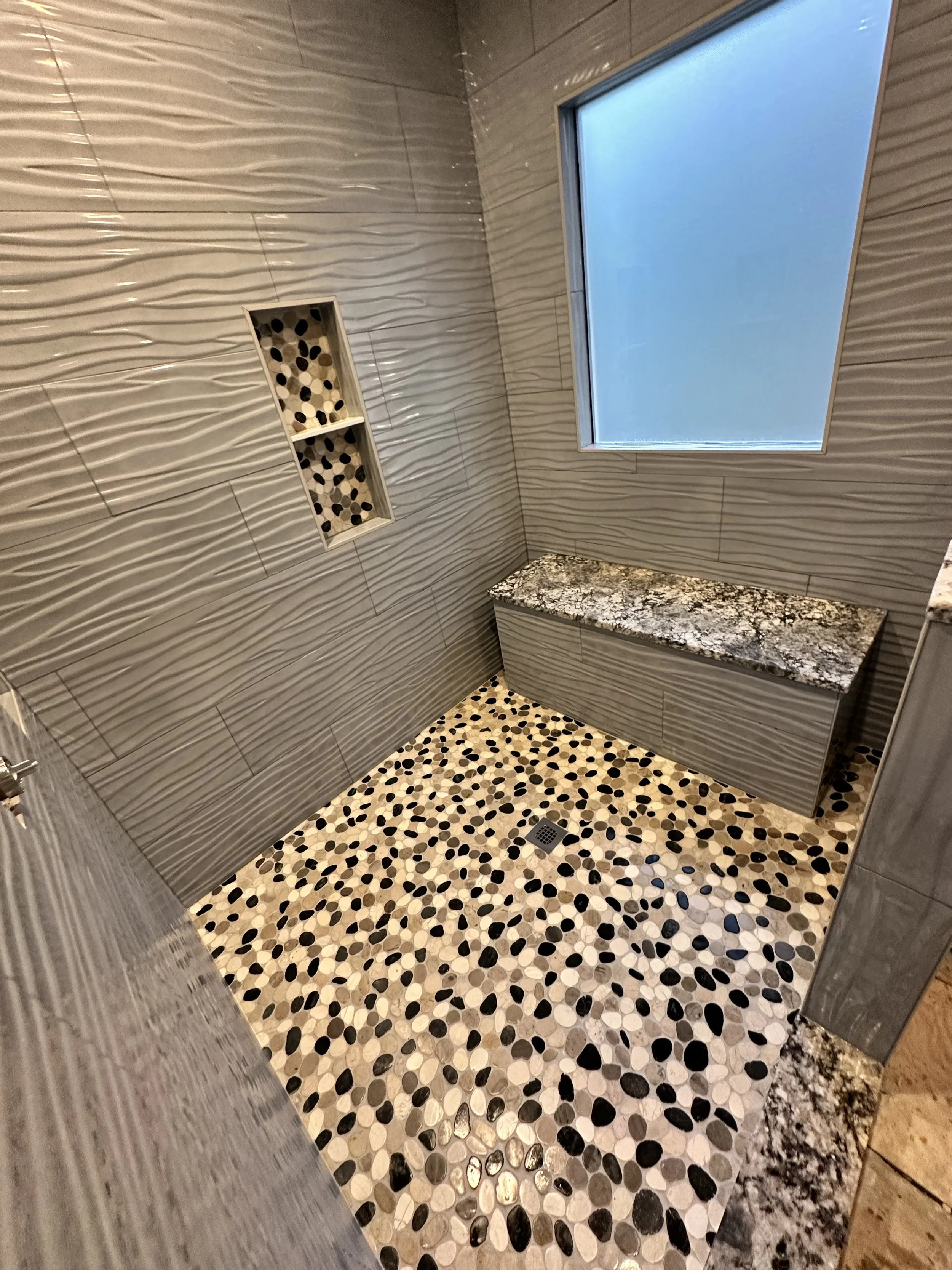 Empty bathroom with textured gray tile walls, pebble-patterned floor, granite bench, and a frosted window.