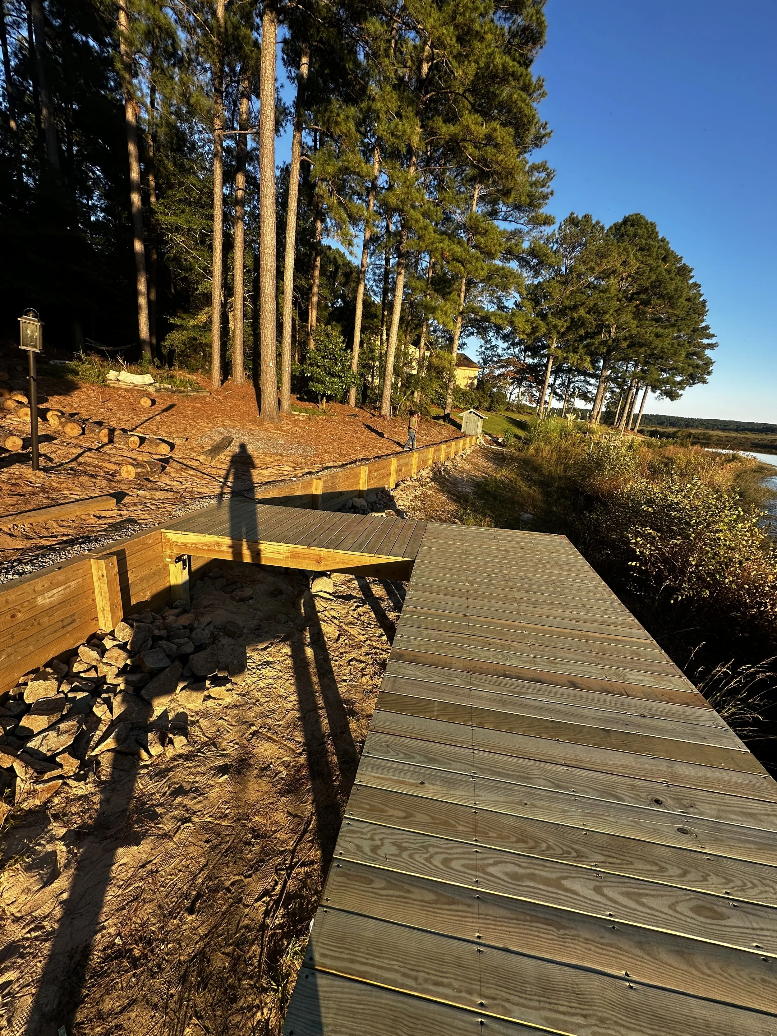 A wooden walkway by a lakeshore with trees, bushes, and houses in the background, during late afternoon or early evening.