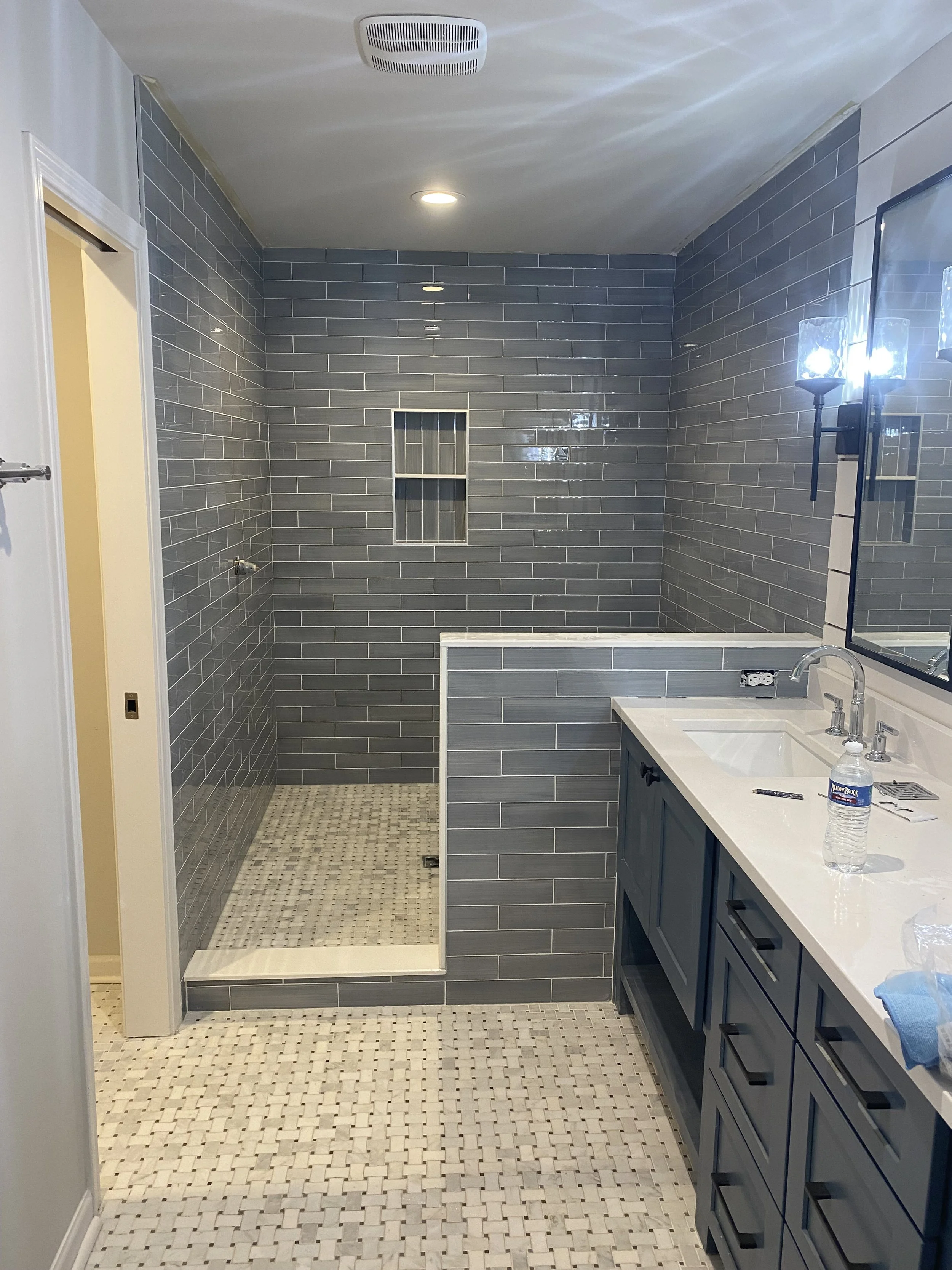 Bathroom with a glass-enclosed shower, gray tiled walls, a white vanity with a sink, a mirror, and a water bottle on the counter.
