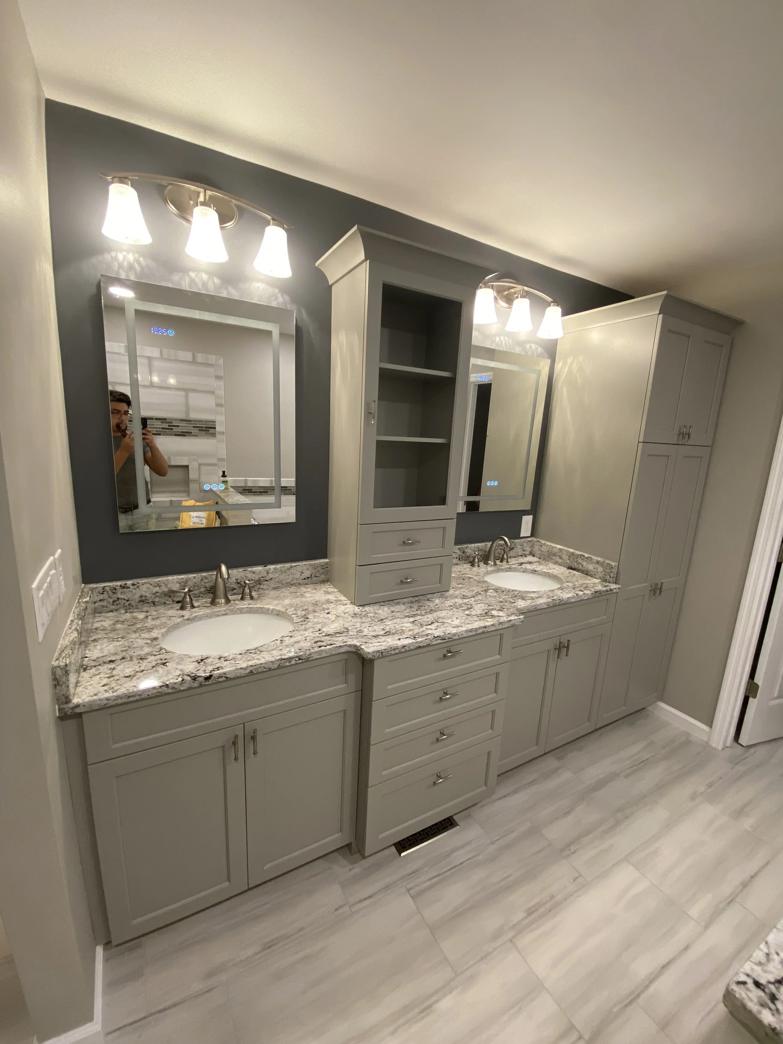 Double bathroom vanity with granite countertops, two sinks, and two mirrors, with wall-mounted light fixtures above each mirror, in a modern bathroom with gray cabinetry and tiled floor.