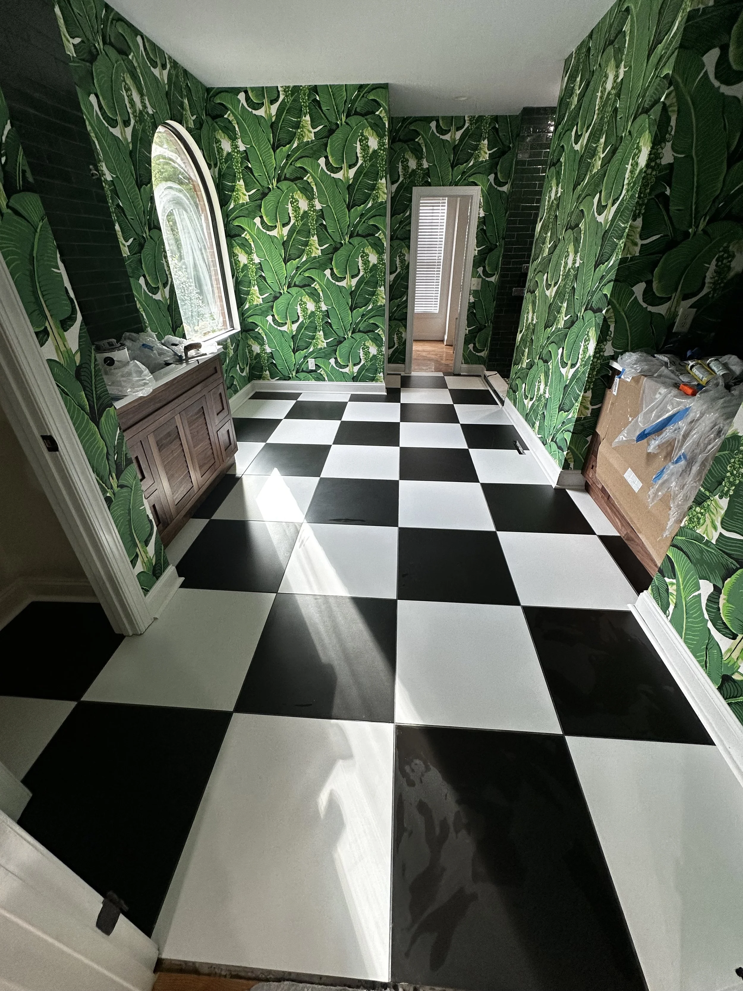 Room with green leafy patterned wallpaper, black and white checkered tile floor, and a small wooden cabinet under a window.