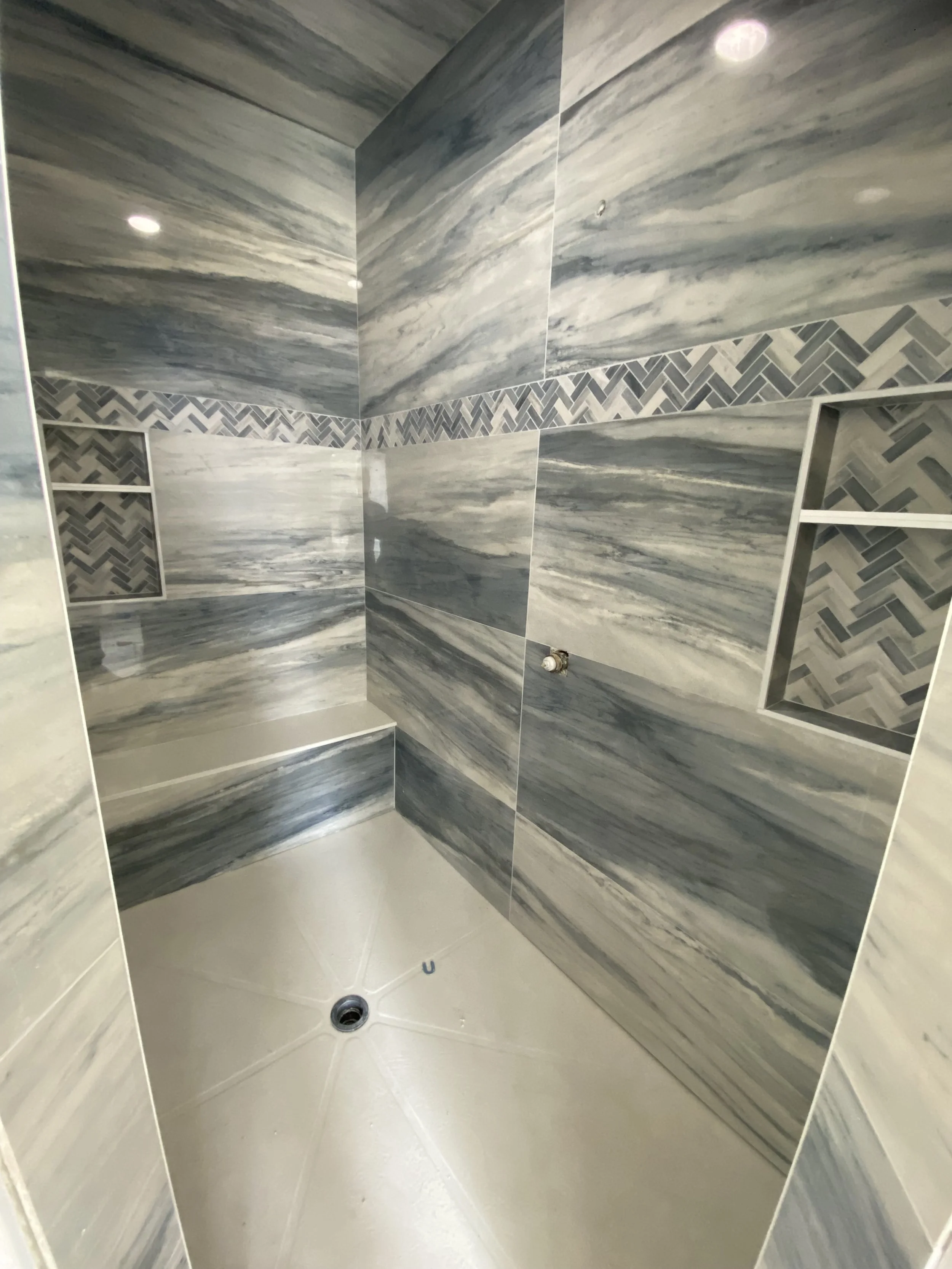 Shower with grey and white marble-like tile walls, built-in shelves, and a beige shower base with a drain.