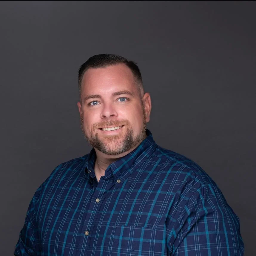 Professional headshot of a smiling man with short brown hair, a beard, wearing a blue plaid shirt against a gray background.