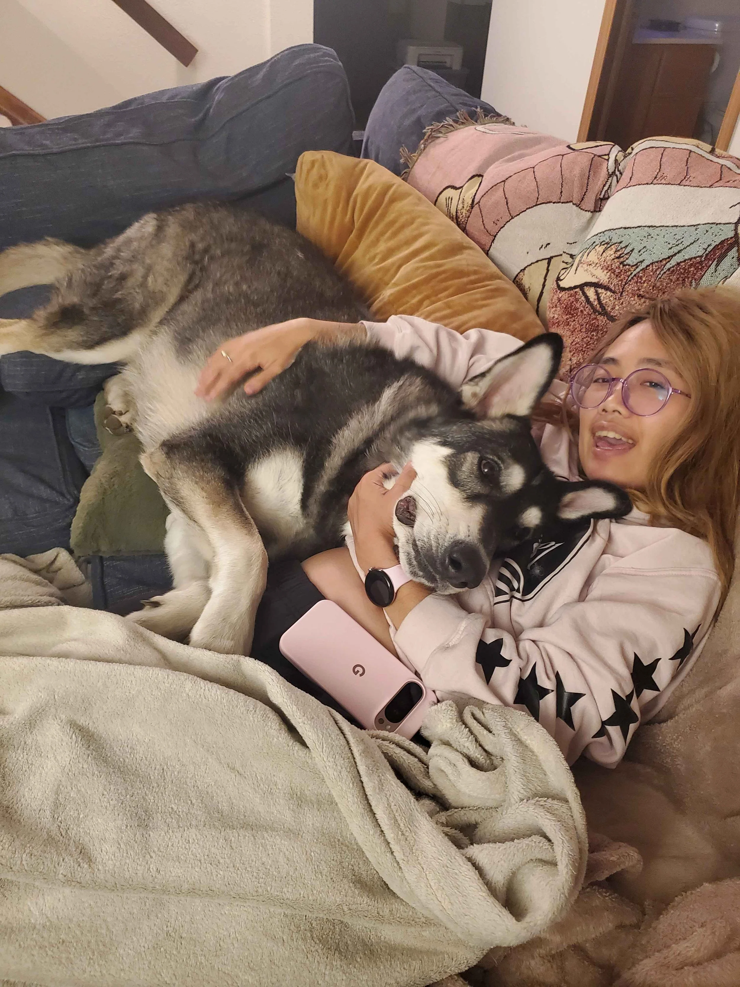 A woman with glasses lying on a couch cuddling with a large husky dog, surrounded by pillows and a blanket.