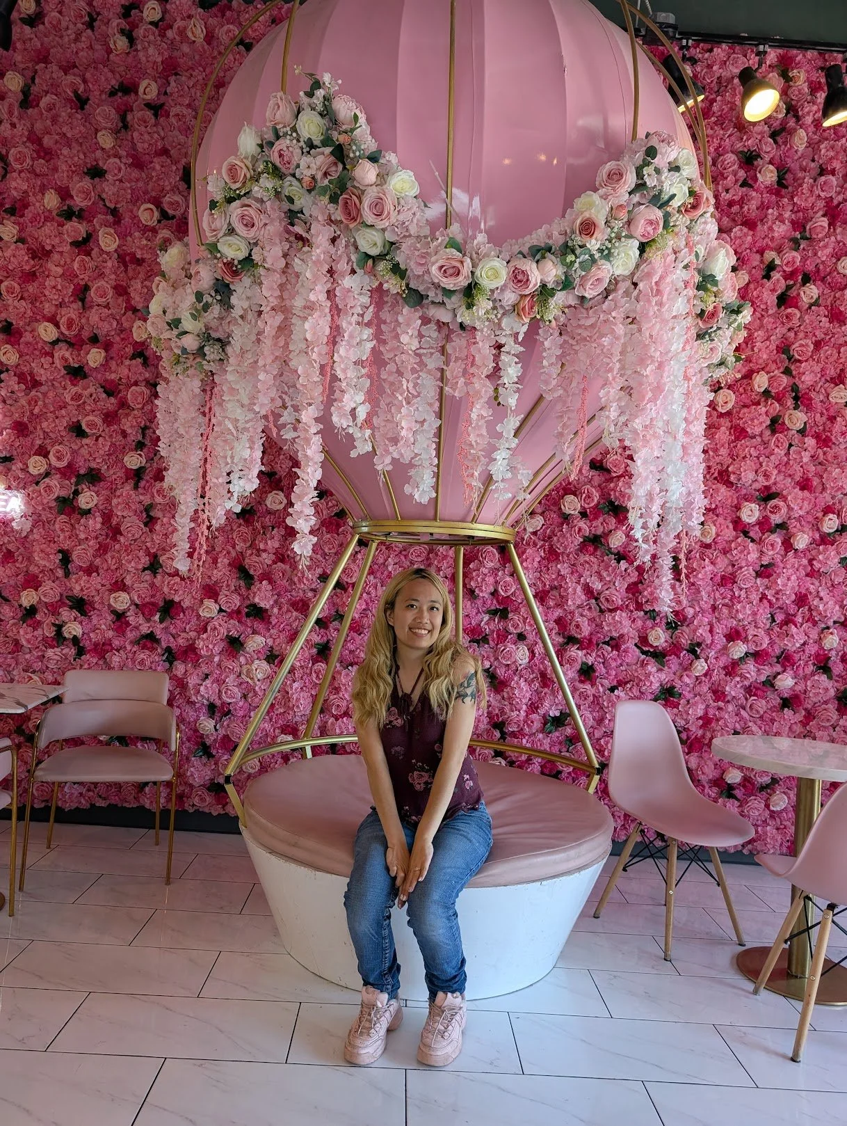 A young woman sits on a pink cushioned chair in front of a wall covered in pink roses. Behind her is a hanging decorative structure resembling a hot air balloon with pink and white flowers, set in a floral-themed room with pink chairs and tables.