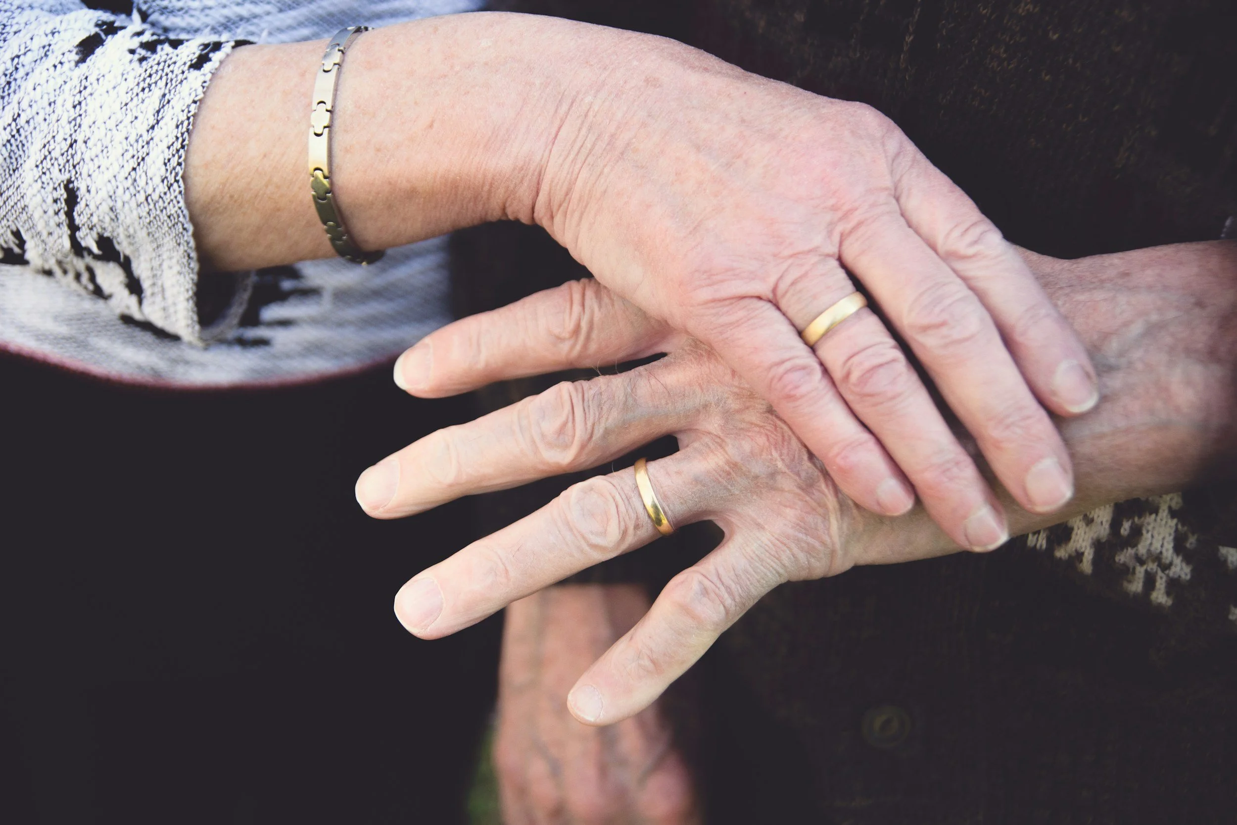 Close-up of two hands with wedding bands, one elderly woman's hand resting on top of an elderly man's hand, outdoors.