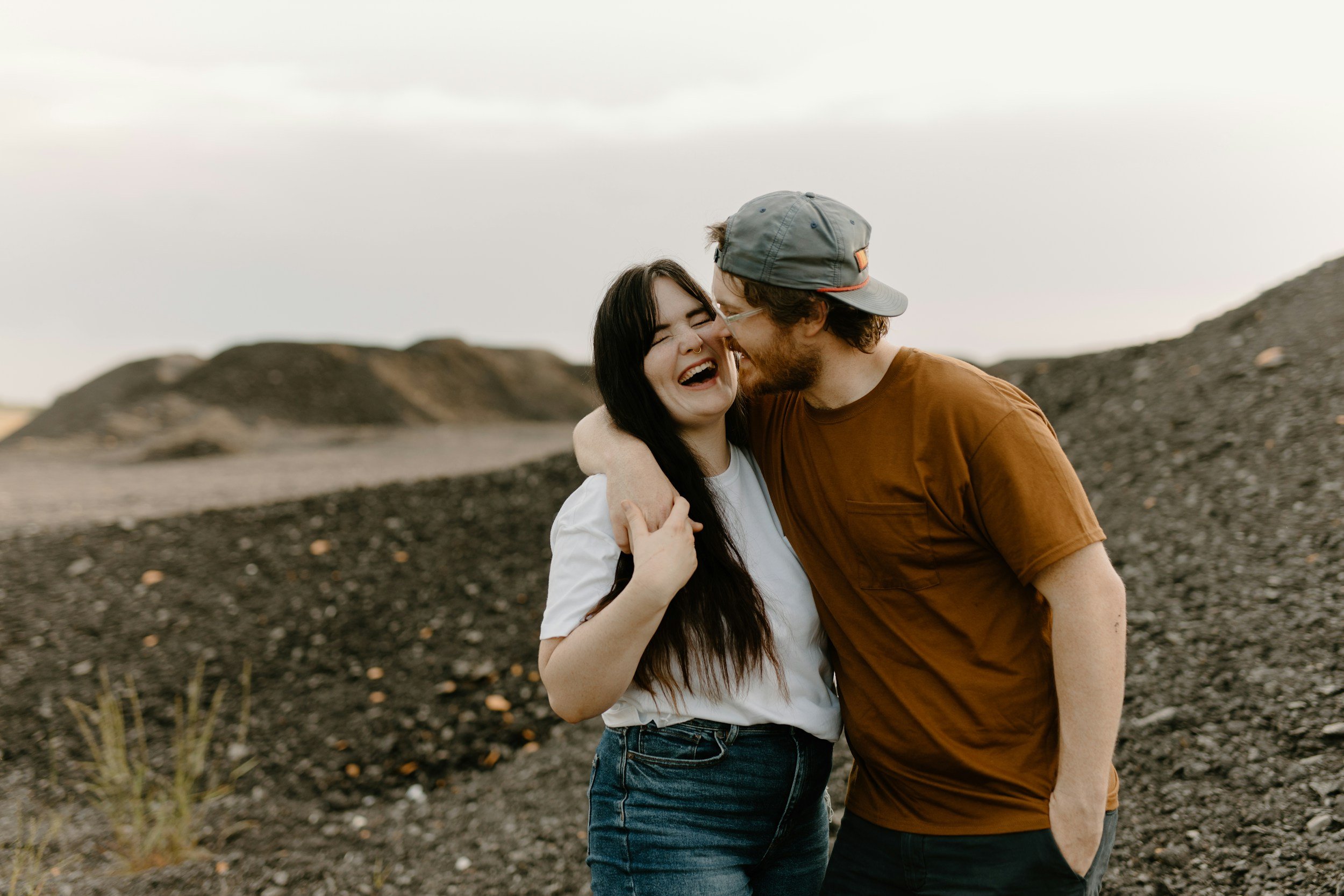 A happy couple laughing and hugging outdoors on a rocky terrain with hills in the background.