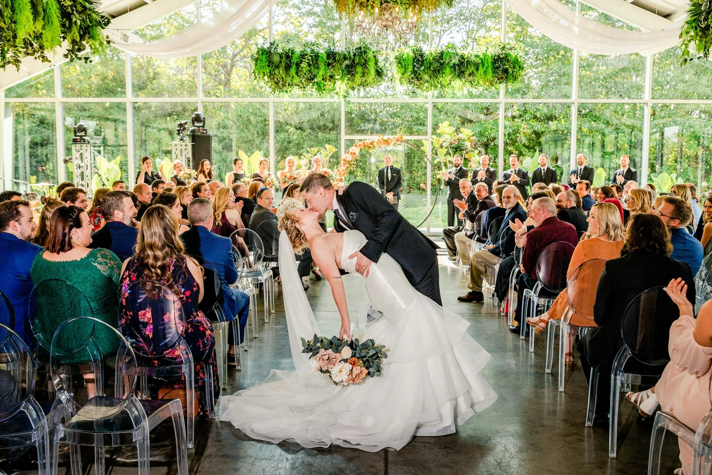 A newlywed couple sharing a kiss during their wedding ceremony, with the bride in a white gown holding a bouquet and the groom in a black suit, surrounded by seated guests and a sunlit glass-enclosed venue with greenery outside.