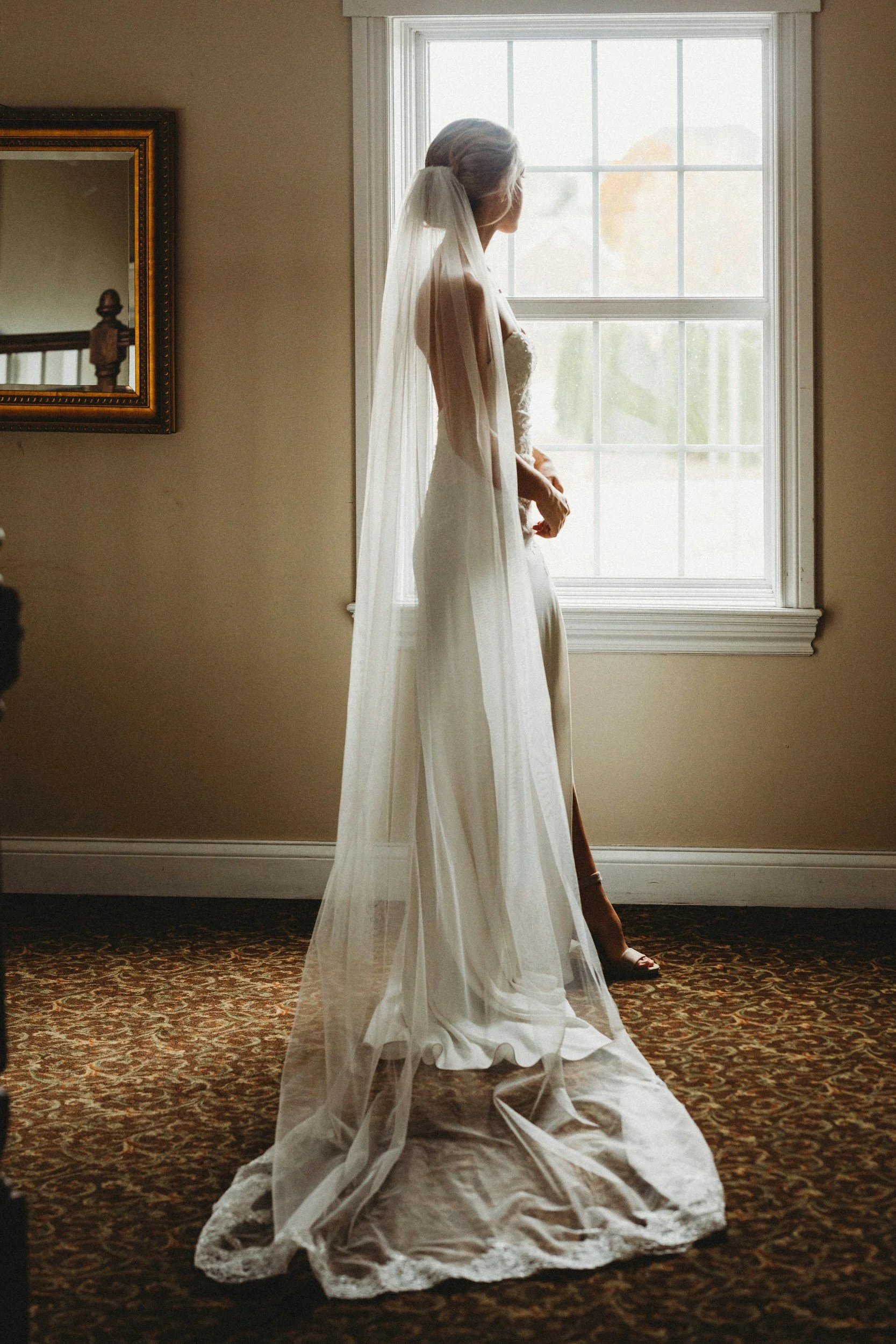 Bride standing by a window in a wedding dress with a long veil, looking outside.
