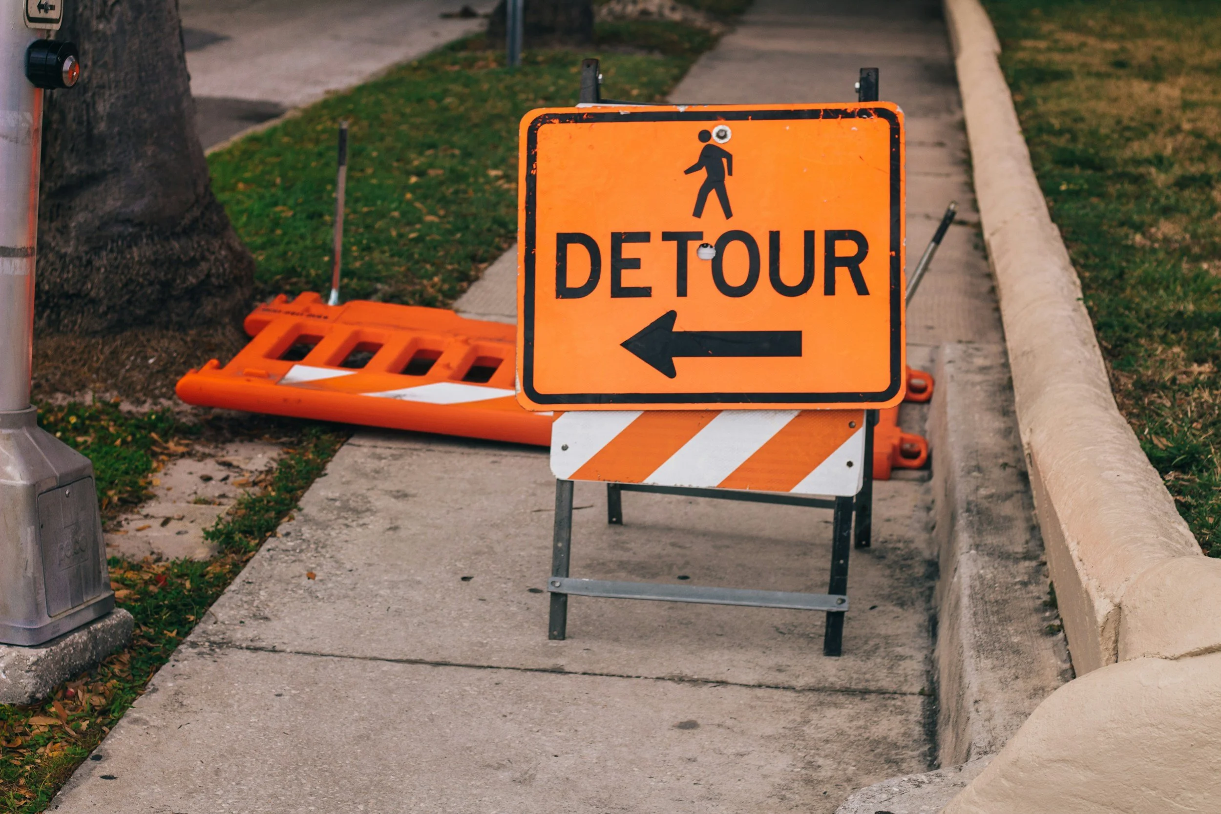 Construction sidewalk with a bright orange "Detour" sign and an arrow pointing left, orange barricades, and equipment nearby.