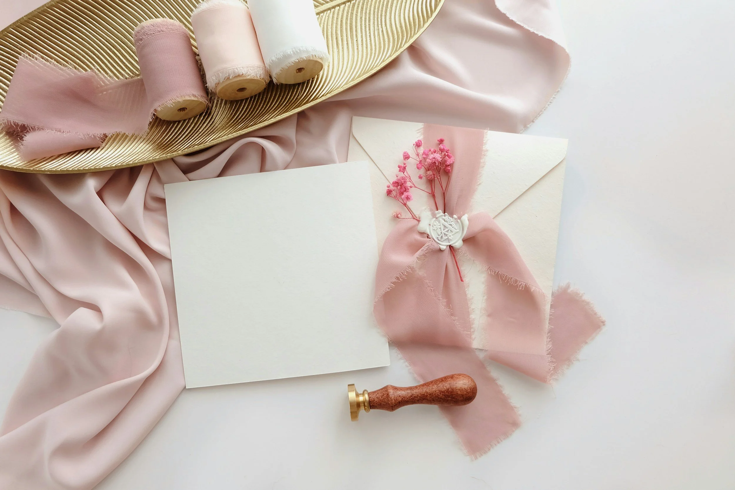 Decorative items for a wedding invitation include pink and white spools of thread, blush pink silk fabric, an envelope with a pink ribbon and small pink flowers, and a wax stamp with a wooden handle on a white background.