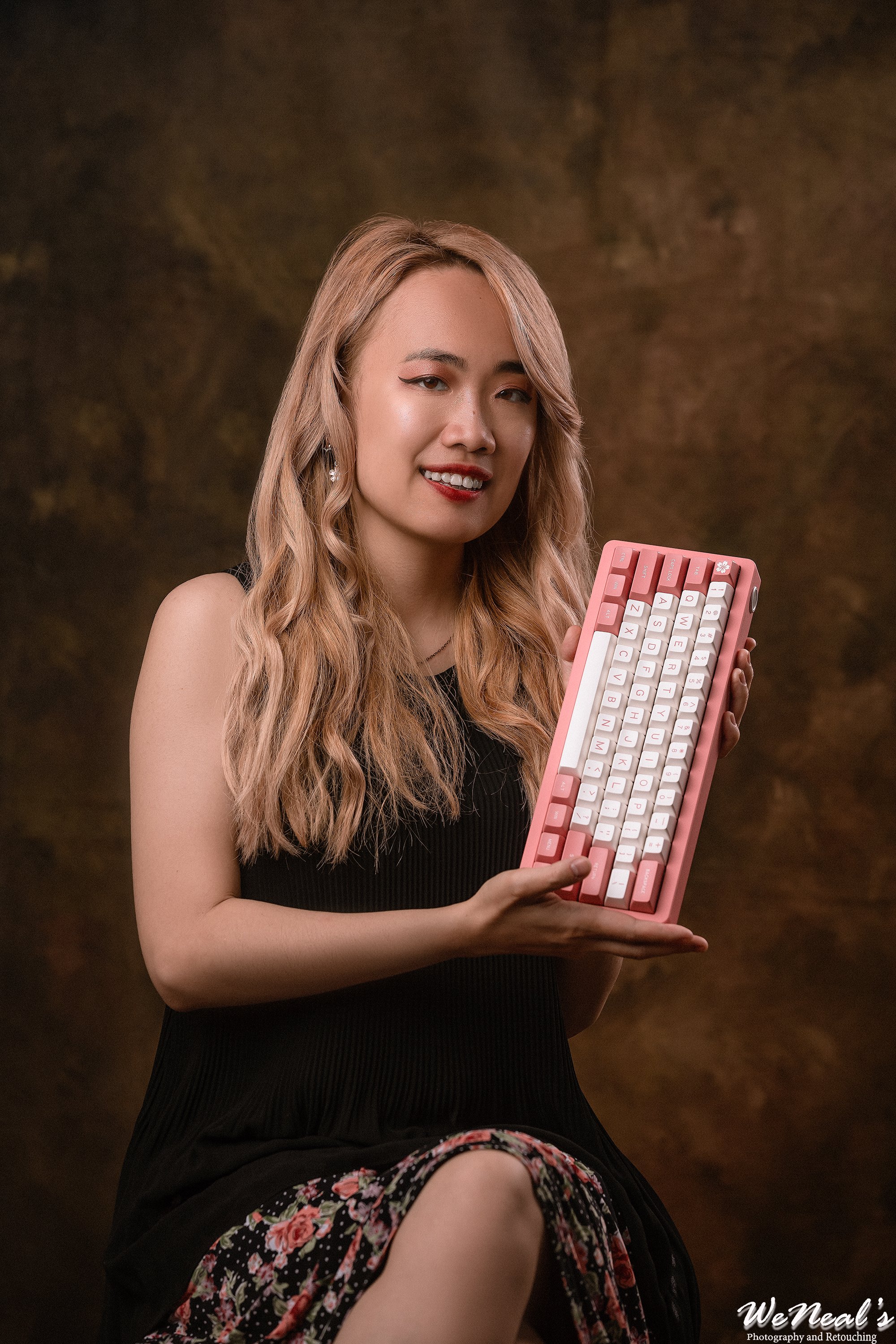 A woman with long blonde wavy hair wearing a black sleeveless top and floral skirt, holding a pink and white keyboard against a brown textured background.