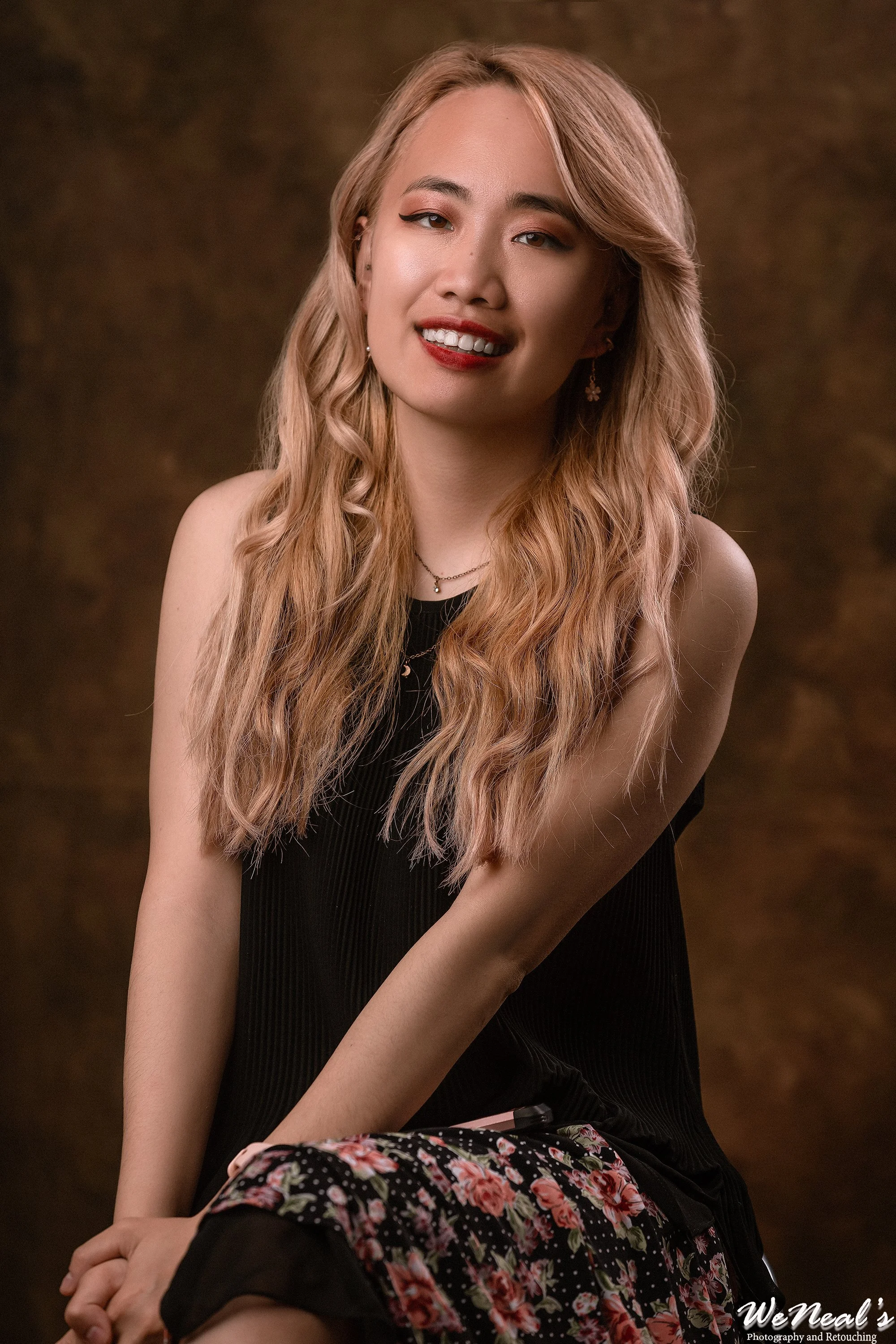 A young woman with long, wavy blonde hair, wearing a black sleeveless top and floral skirt, smiling while posing for a portrait against a brown textured background.