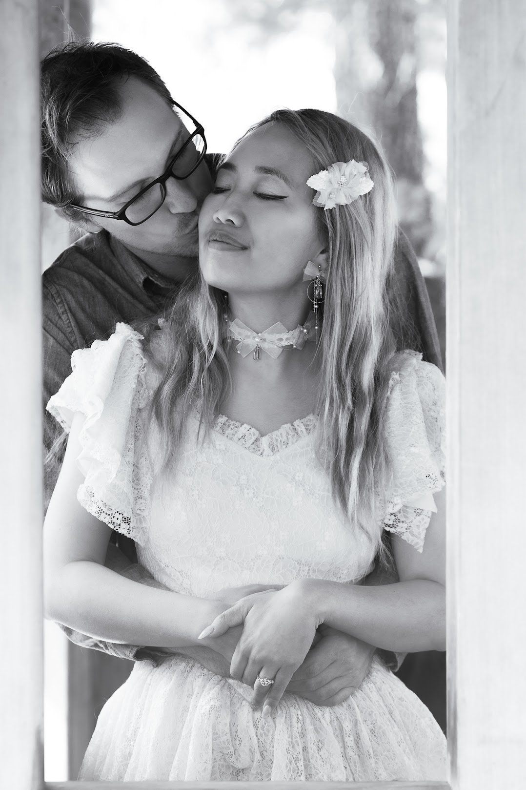 A couple embraces through a window; the man gives a kiss on the woman's cheek as she smiles with her eyes closed, wearing a white lace dress, floral hair accessory, and jewelry.