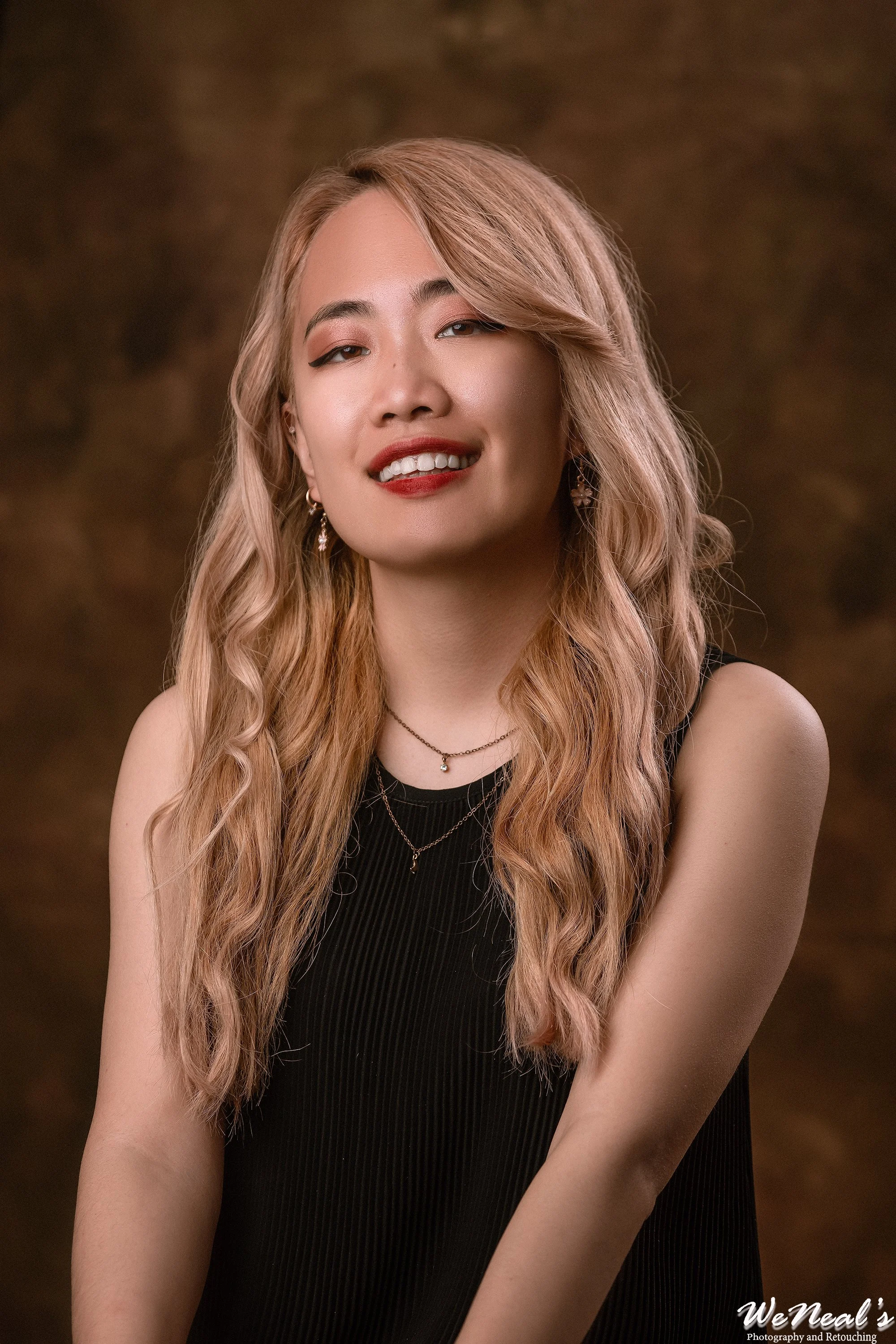 Portrait of a woman with long, wavy, blonde hair, wearing a black sleeveless top and jewelry, smiling in front of a brown background.