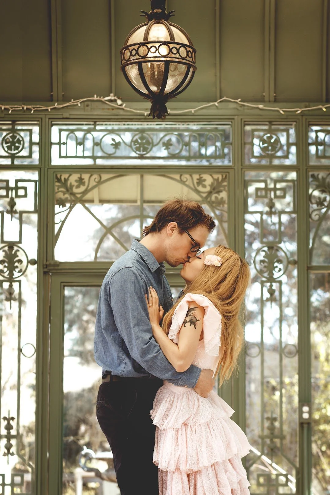 A couple sharing a kiss indoors with decorative green ironwork and large windows in the background.
