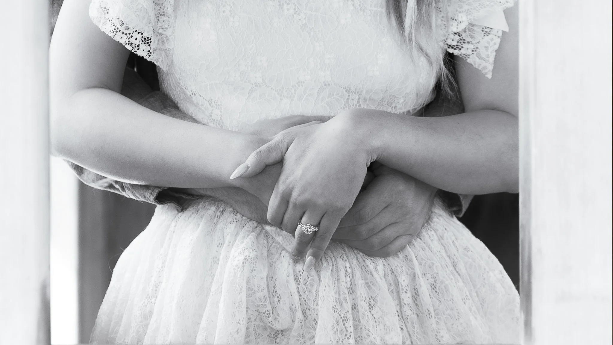 A woman in a lace dress with her hands crossed, showing a wedding ring on her finger.