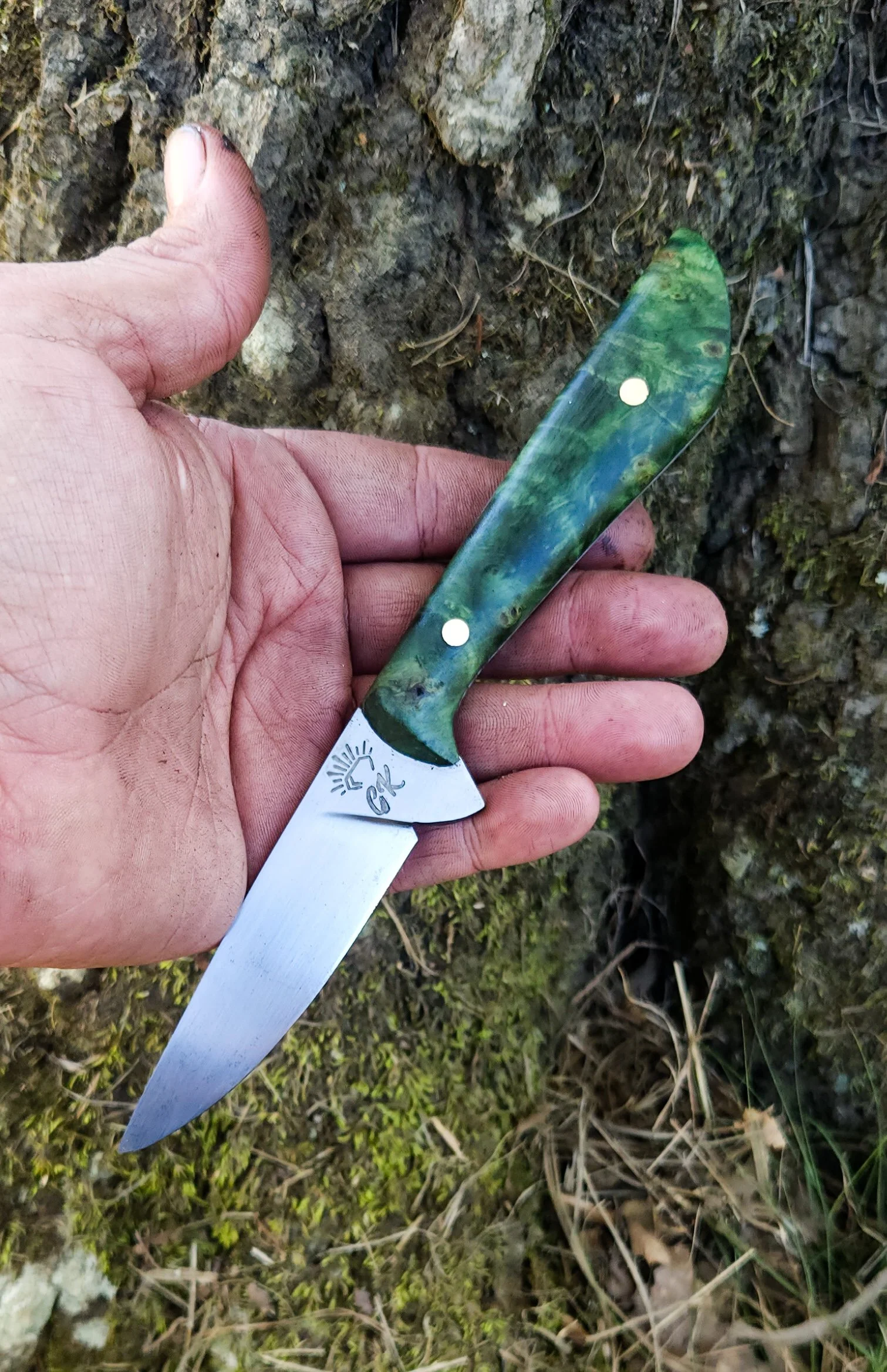 A person's hand holding a green and blue patterned folding pocket knife with a shiny blade, outdoors on the ground covered with moss, small rocks, and twigs.