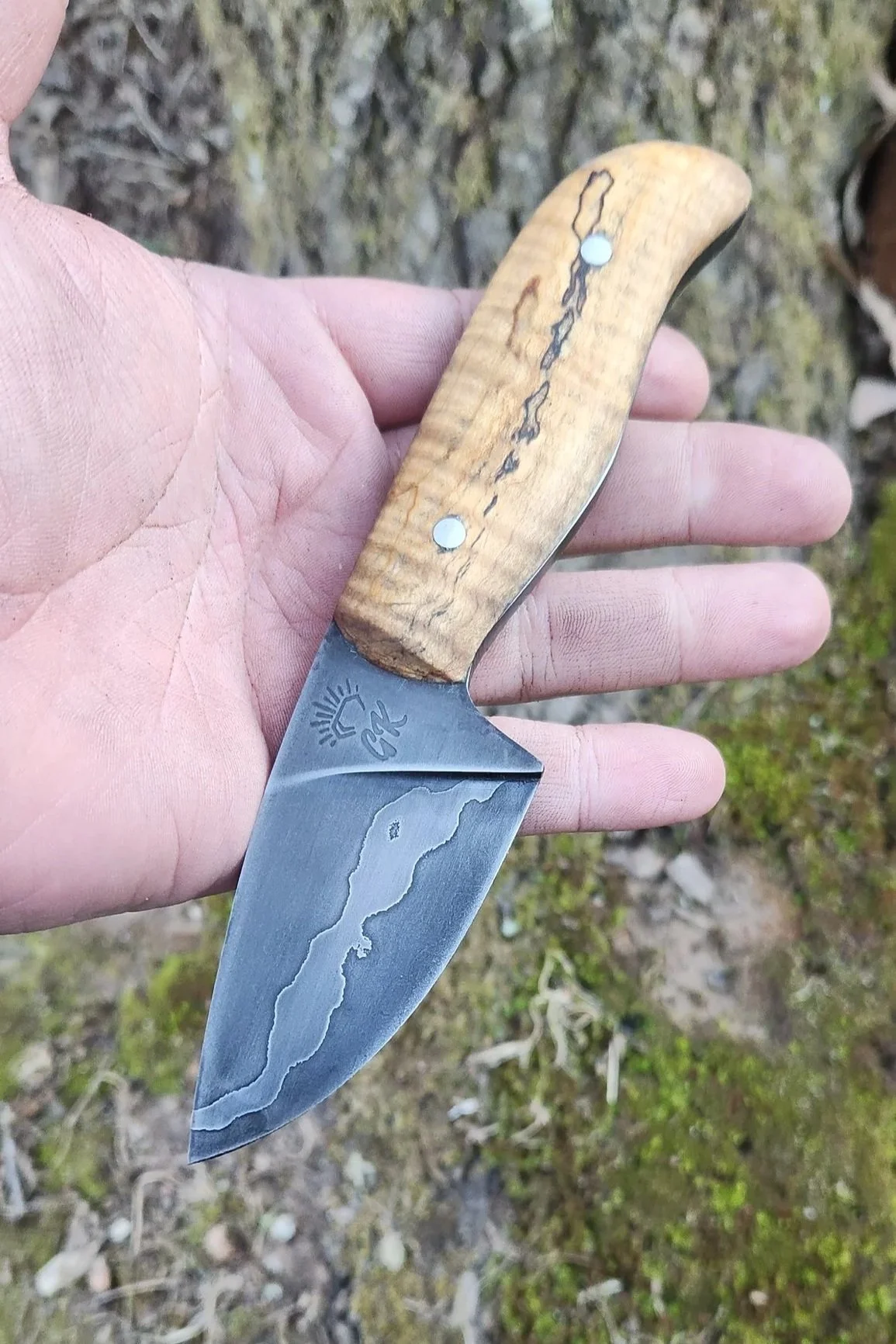 A hand holding a small fixed-blade knife with a wooden handle and a Damascus steel blade with a pattern, outdoors on mossy ground.