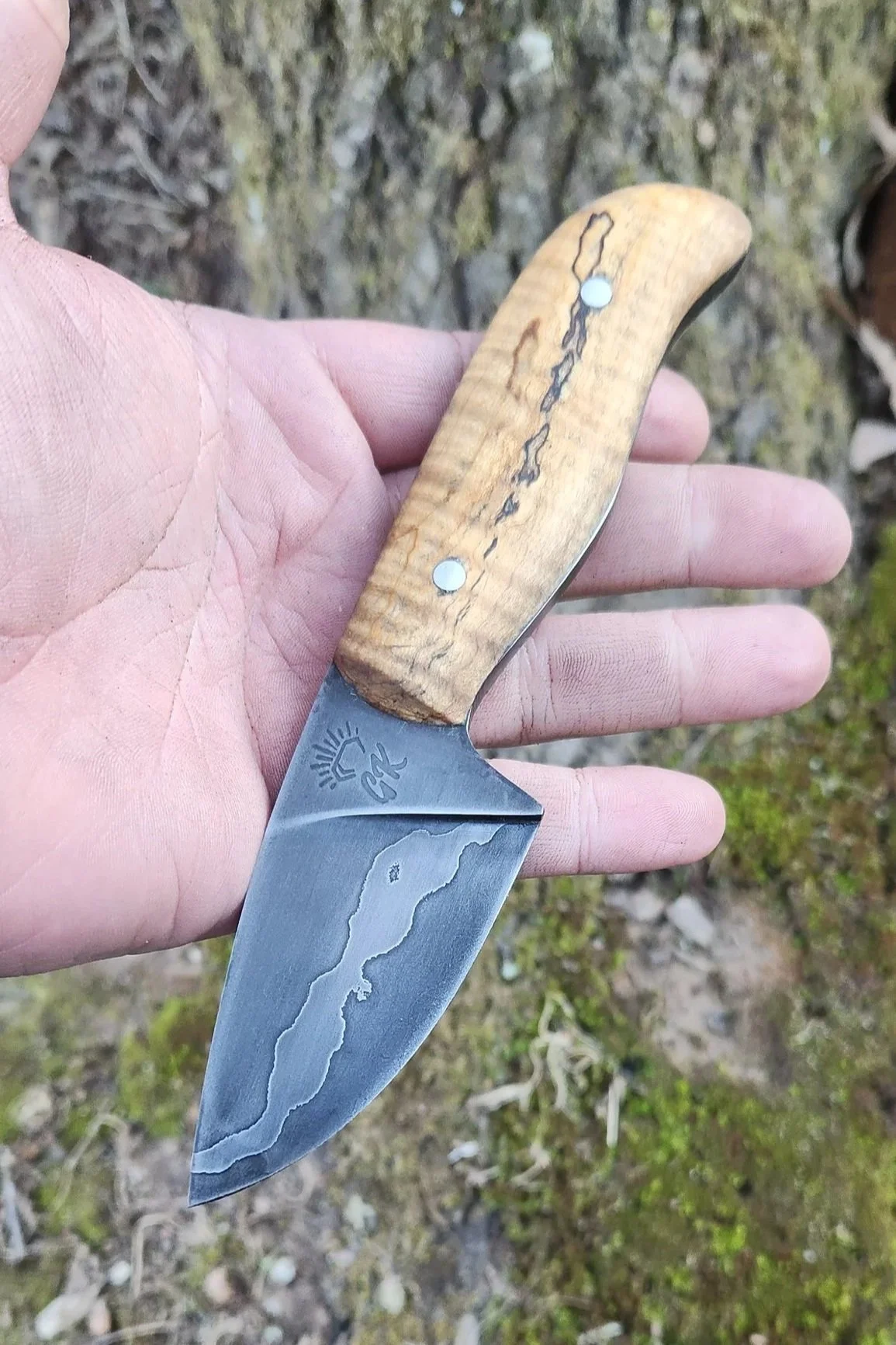A hand holding a small handmade knife with a wooden handle and a Damascus steel blade, outdoors on the ground with moss and leaves.
