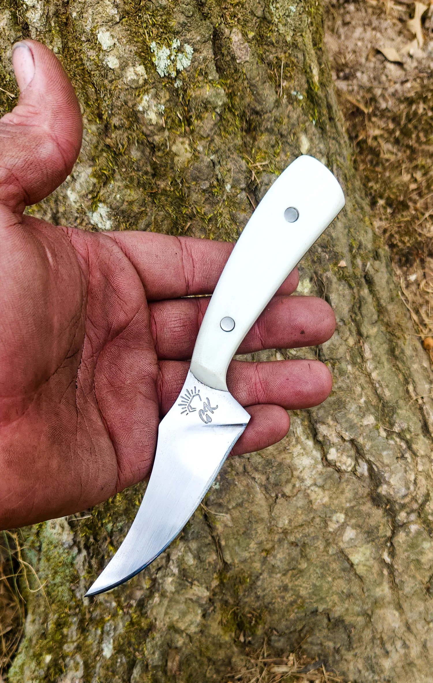 A person holding a small folding knife with a curved blade and white handle, outdoors near tree bark.