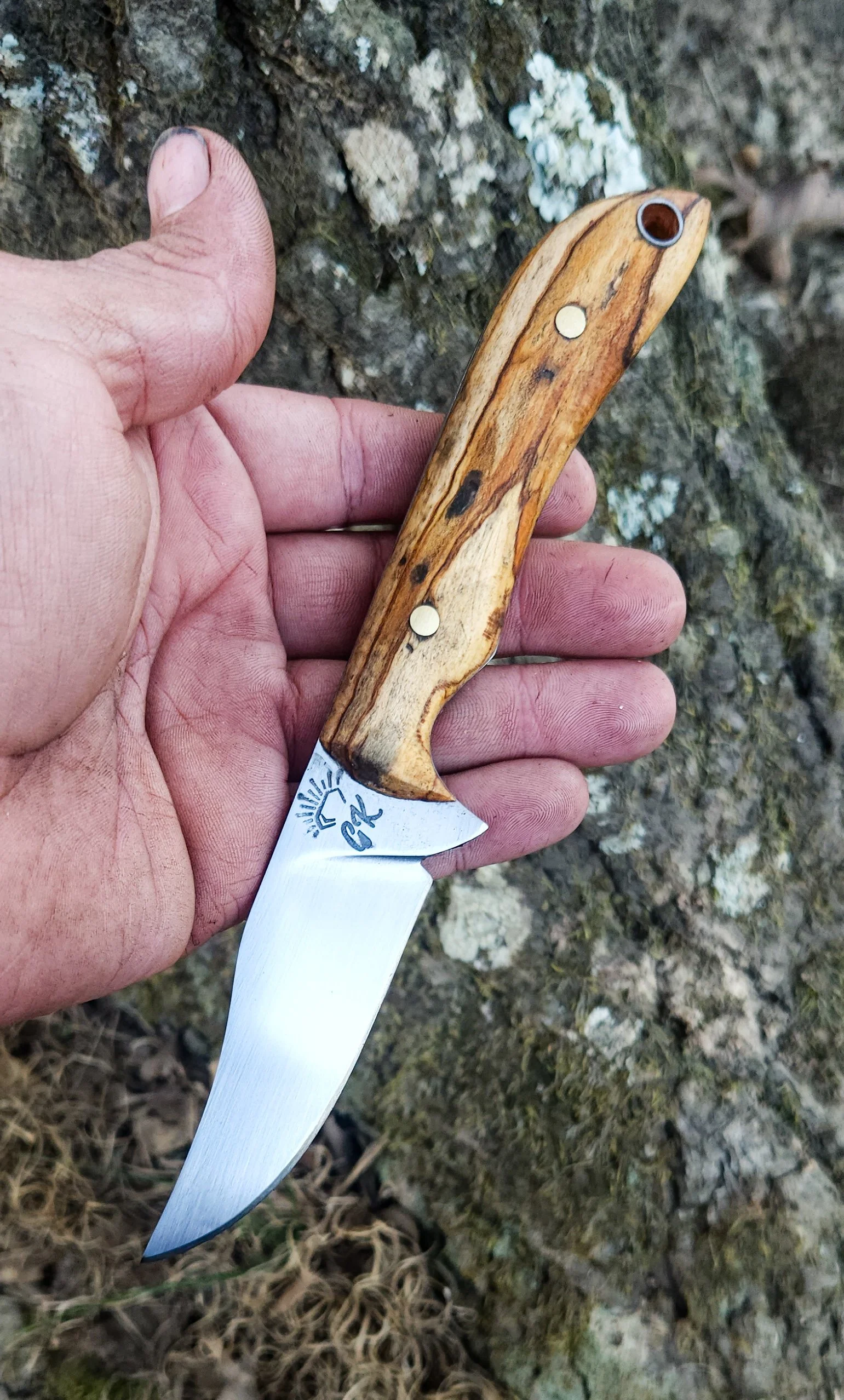 A person's hand holding a small fixed-blade knife with a wooden handle and a sharp, curved metal blade, held against a natural outdoor background with rocks and moss.