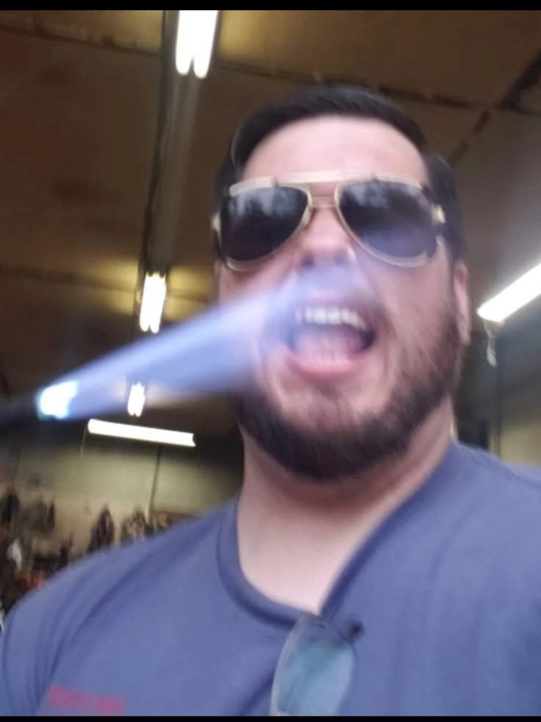 A man with dark hair, a beard, and sunglasses, exhaling smoke in an indoor setting with fluorescent lighting.