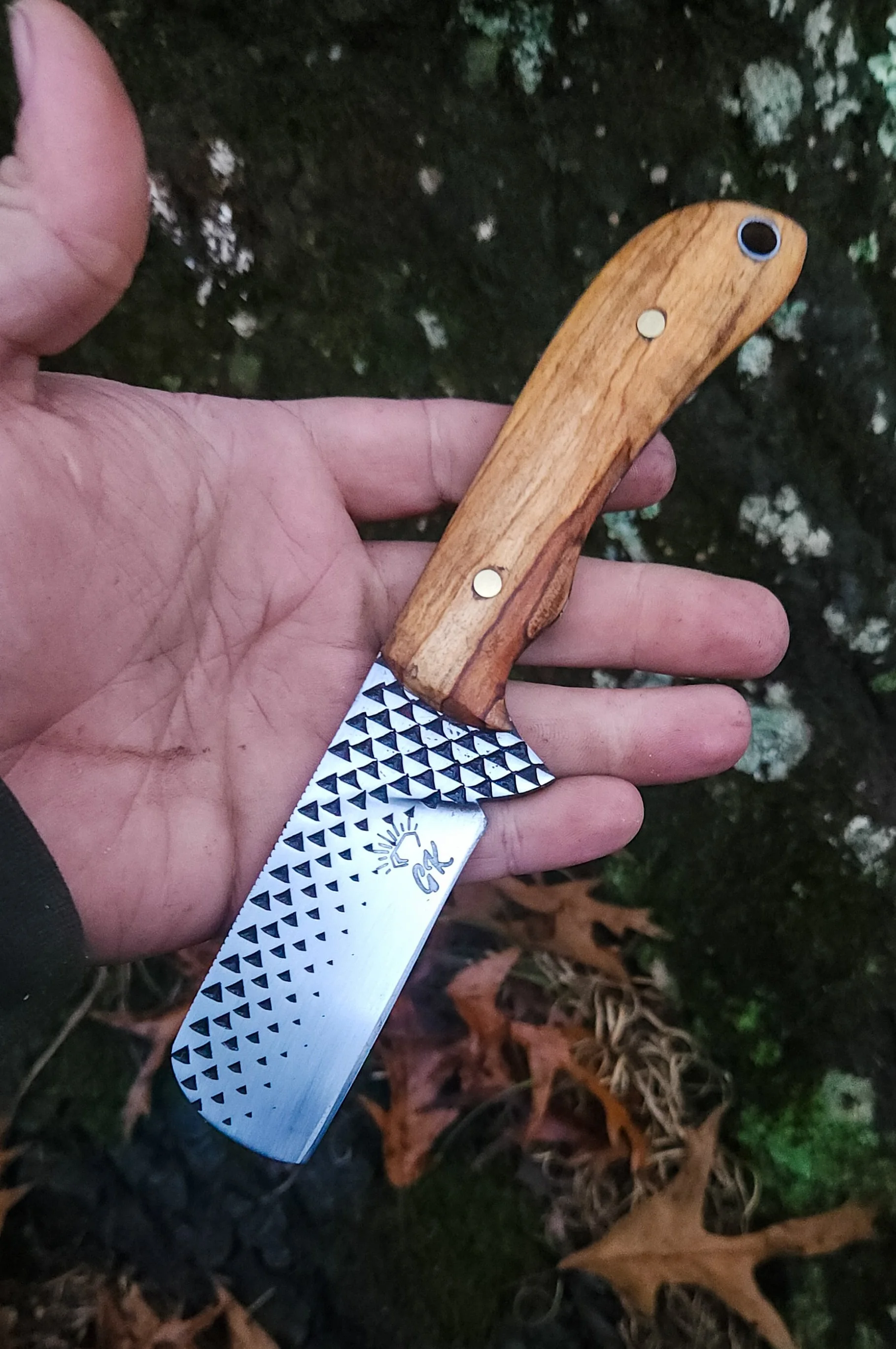 A person holding a kitchen knife with a wooden handle and a patterned stainless steel blade, outdoors against a background of moss and fallen leaves.