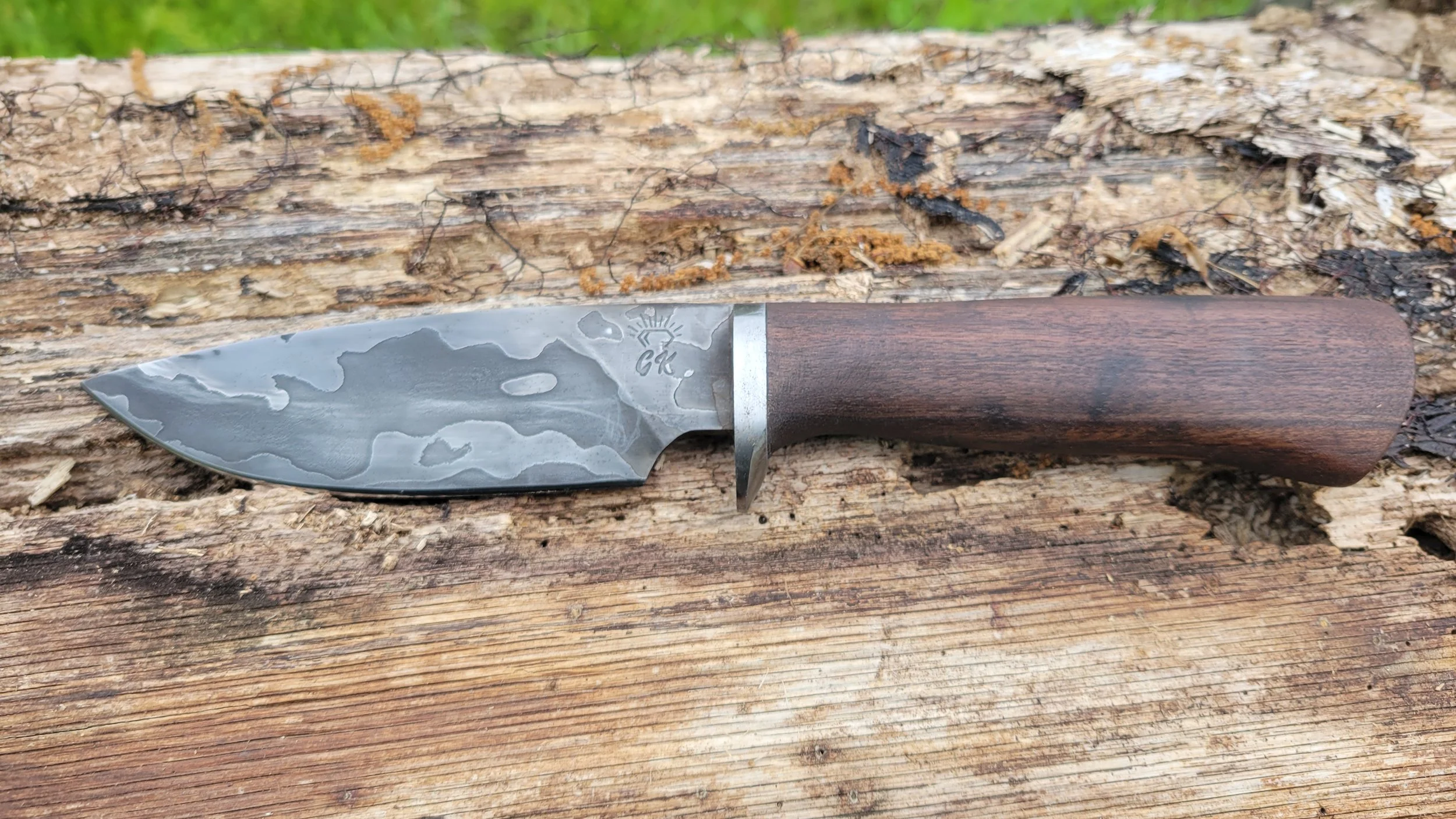A hunting or utility knife with a Damascus steel blade and a wooden handle, resting on a weathered wooden surface outdoors.