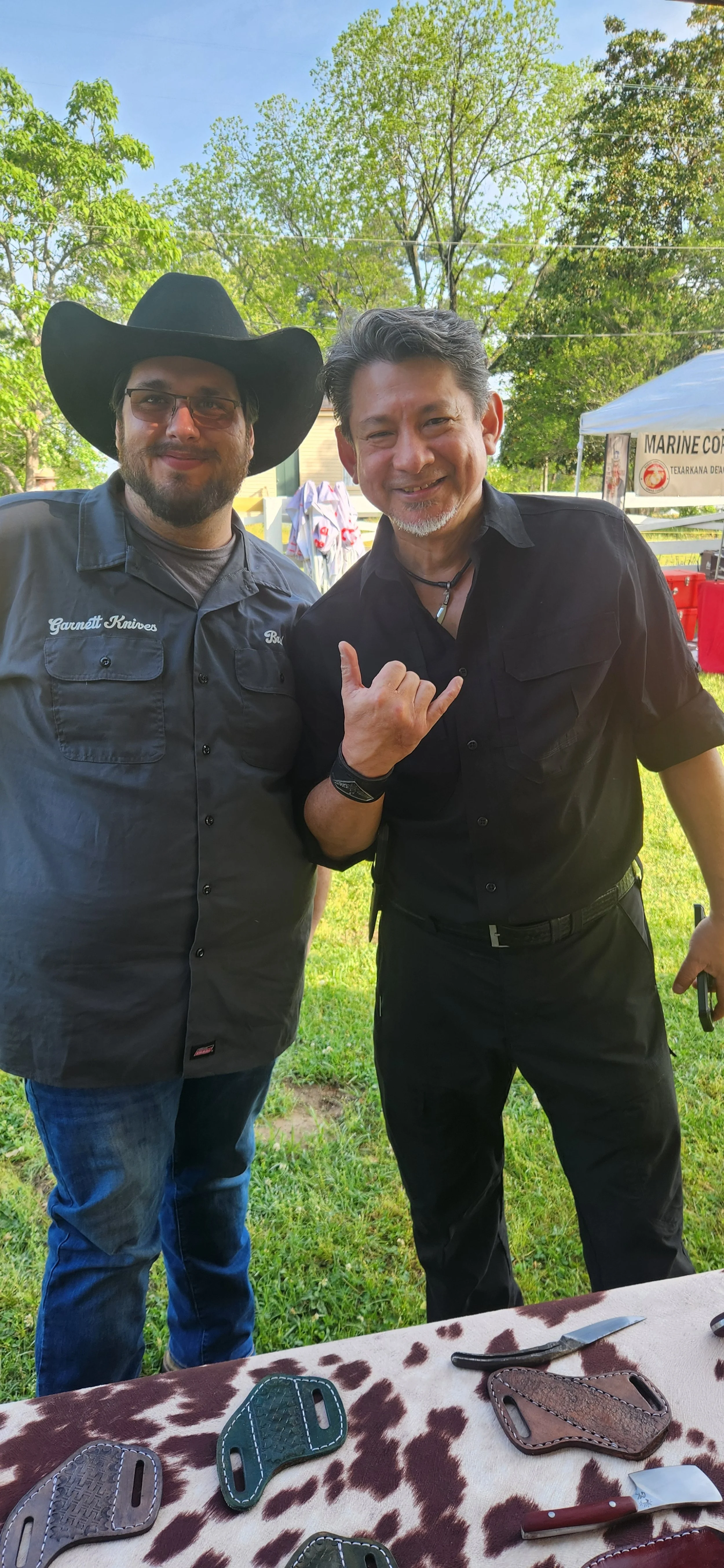 Two men standing outdoors, smiling at the camera. One is wearing a large black cowboy hat and sunglasses, and a gray shirt with patches that say Garnett Knives. The other man is wearing a black shirt, a necklace, and is making a shaka hand gesture. There is a table with knives and leather sheaths in the foreground, and trees and a tent in the background.