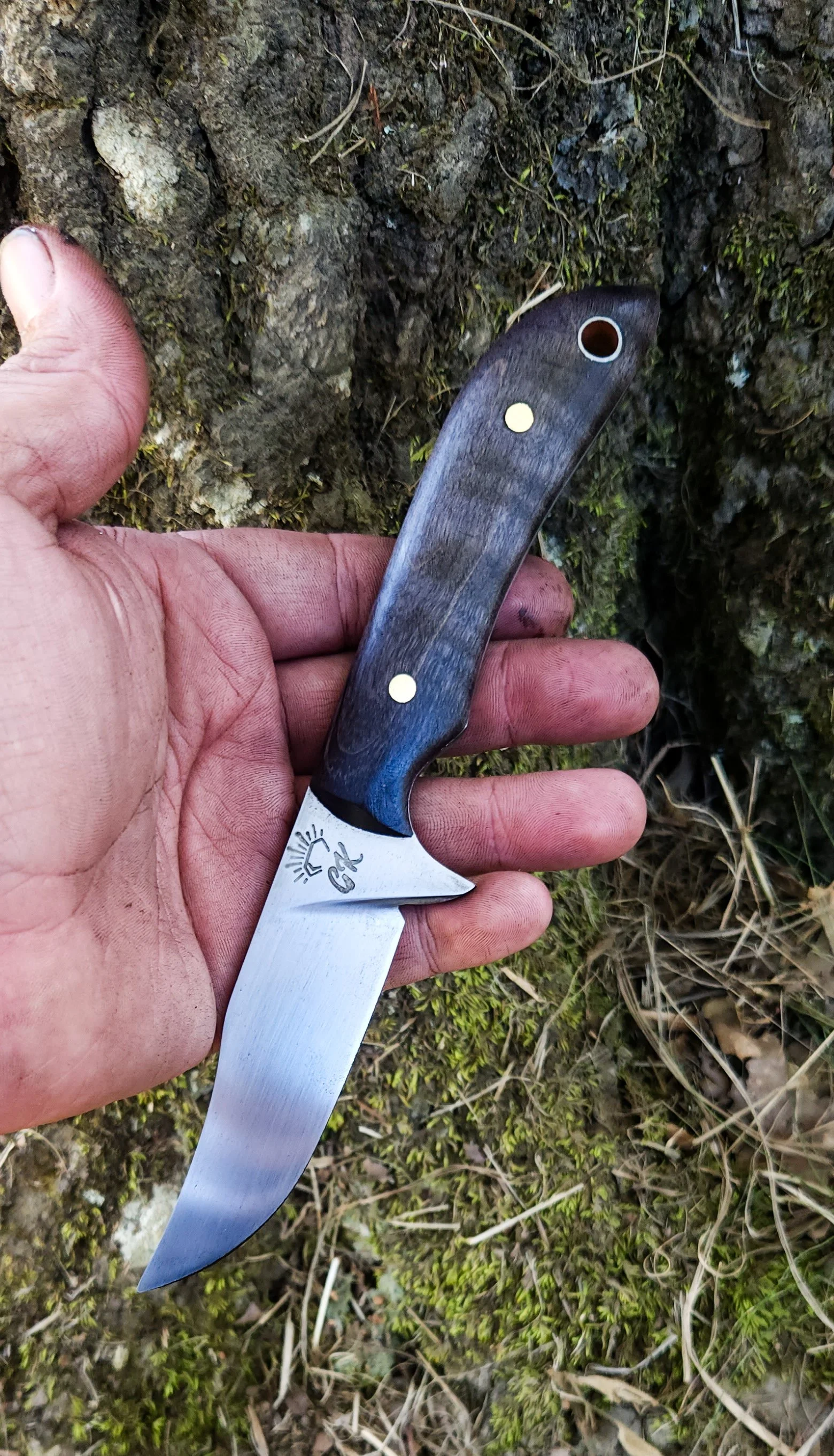 A person's hand holding a small folding knife with a wooden handle and a stainless steel blade outdoors on the ground covered in dirt and moss.