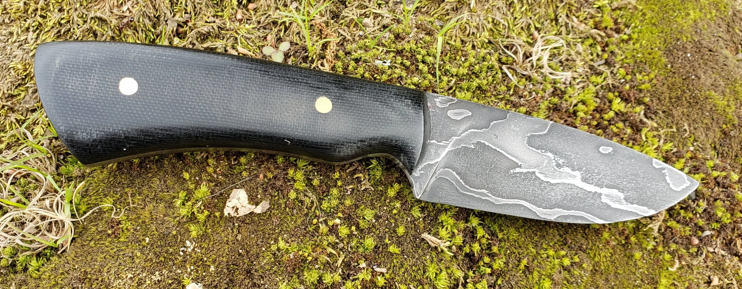 A black Damascus steel knife with a black handle resting on mossy ground with small green plants.