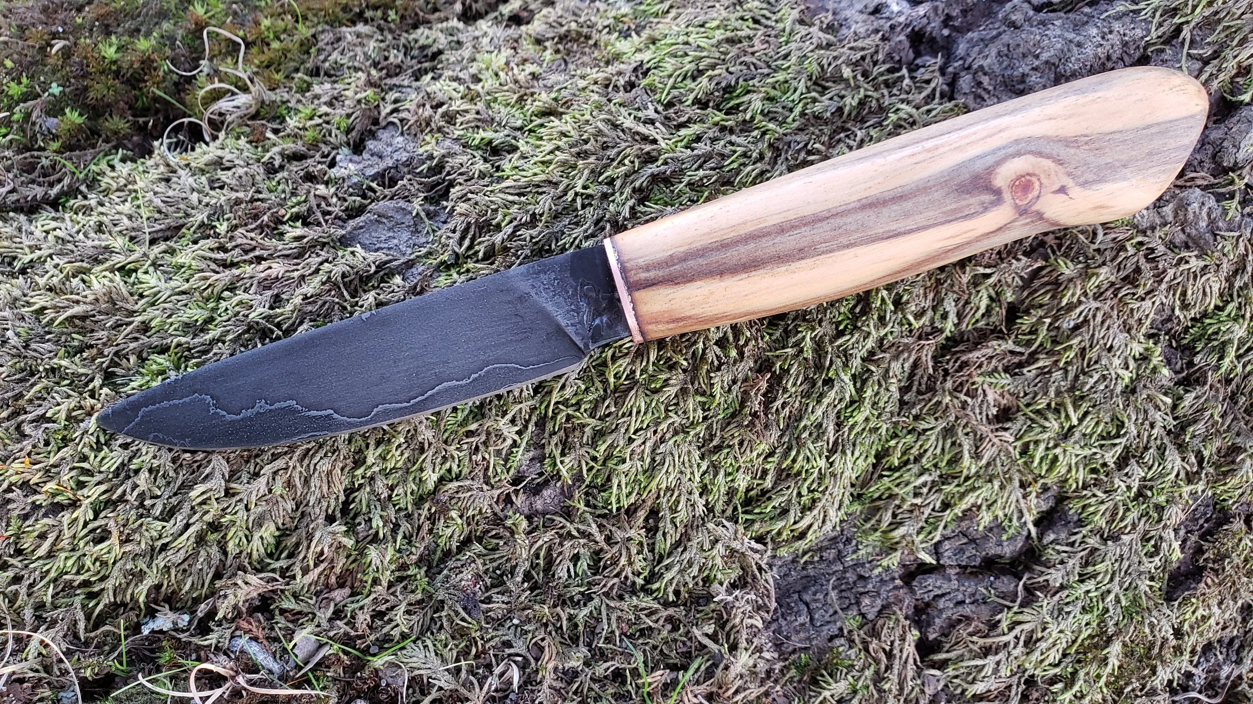 A black-handled knife with a wooden grip lying on mossy ground.