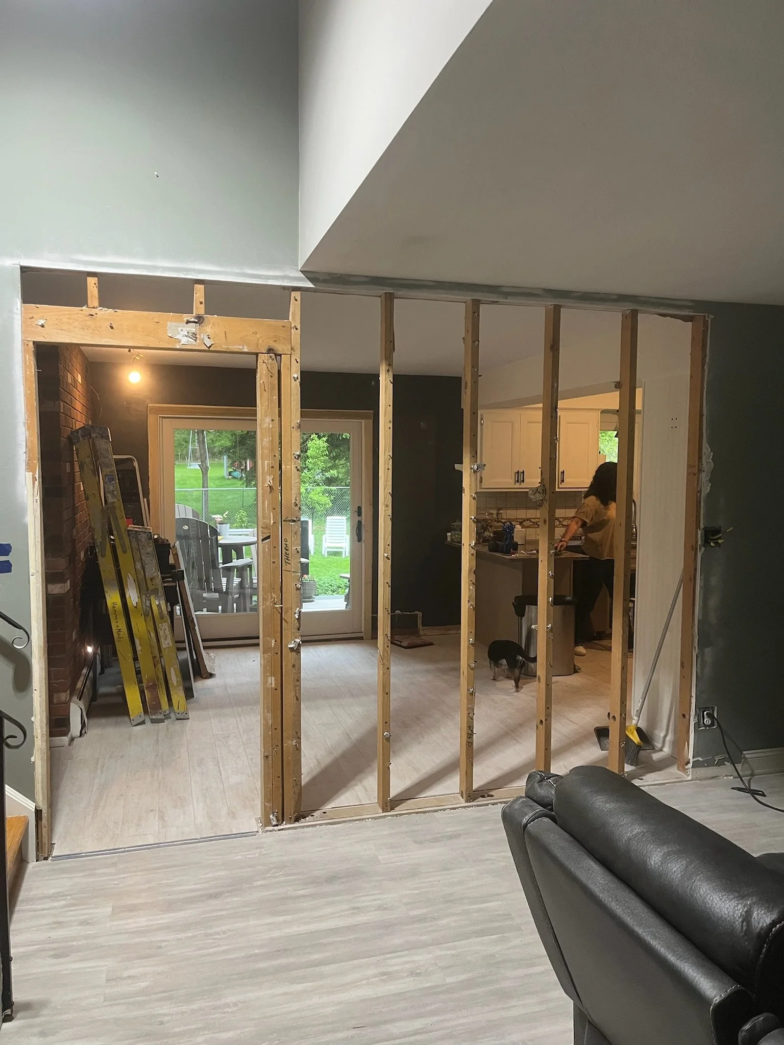 Interior view of a home undergoing renovation, showing framing for a wall with exposed wooden studs, a sliding glass door leading to a backyard, a woman working in the kitchen, and construction tools and equipment near the wall.
