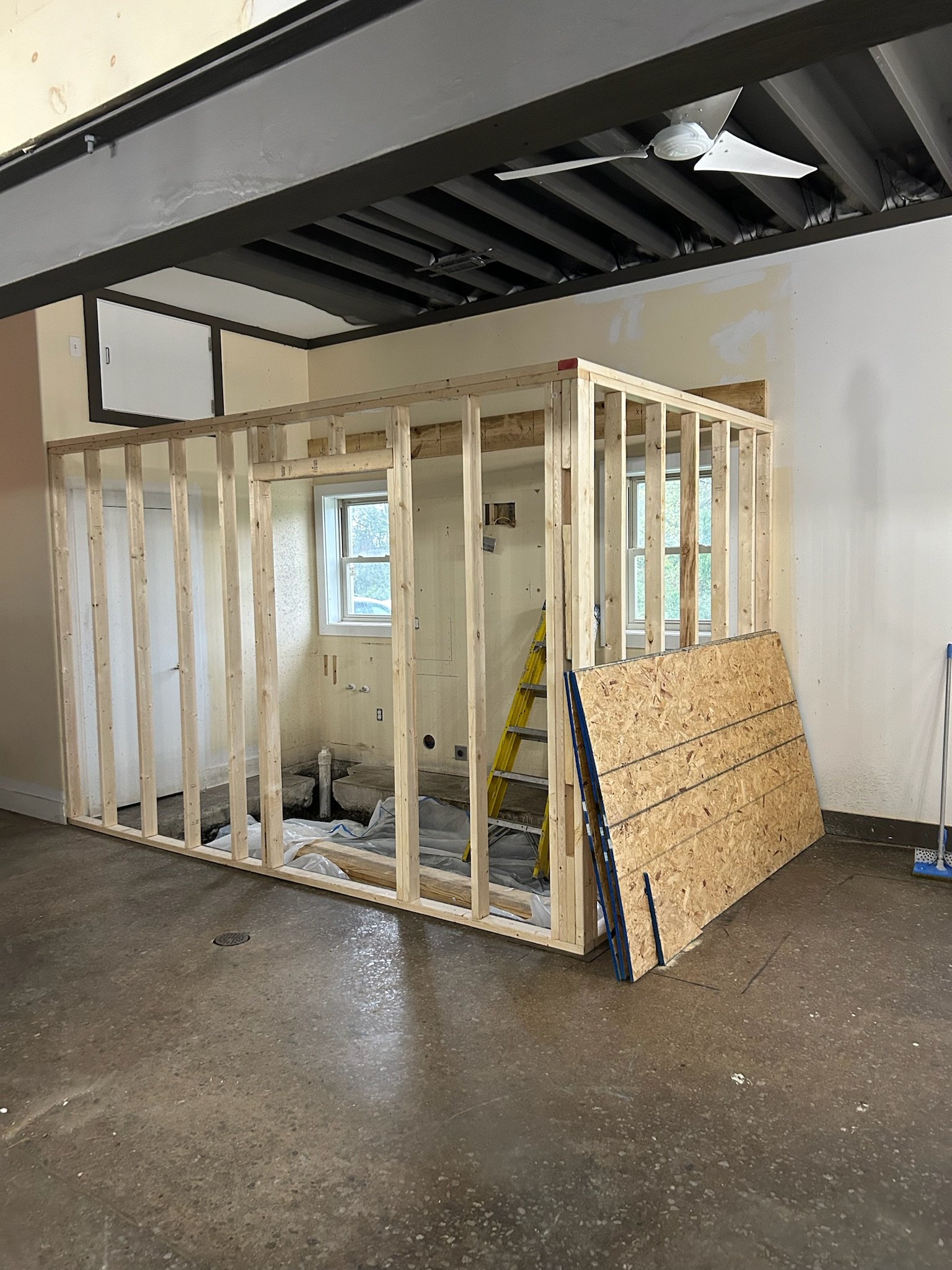 Construction in progress of a small room framed with wooden studs inside a larger unfinished room, with a ladder and a window visible.