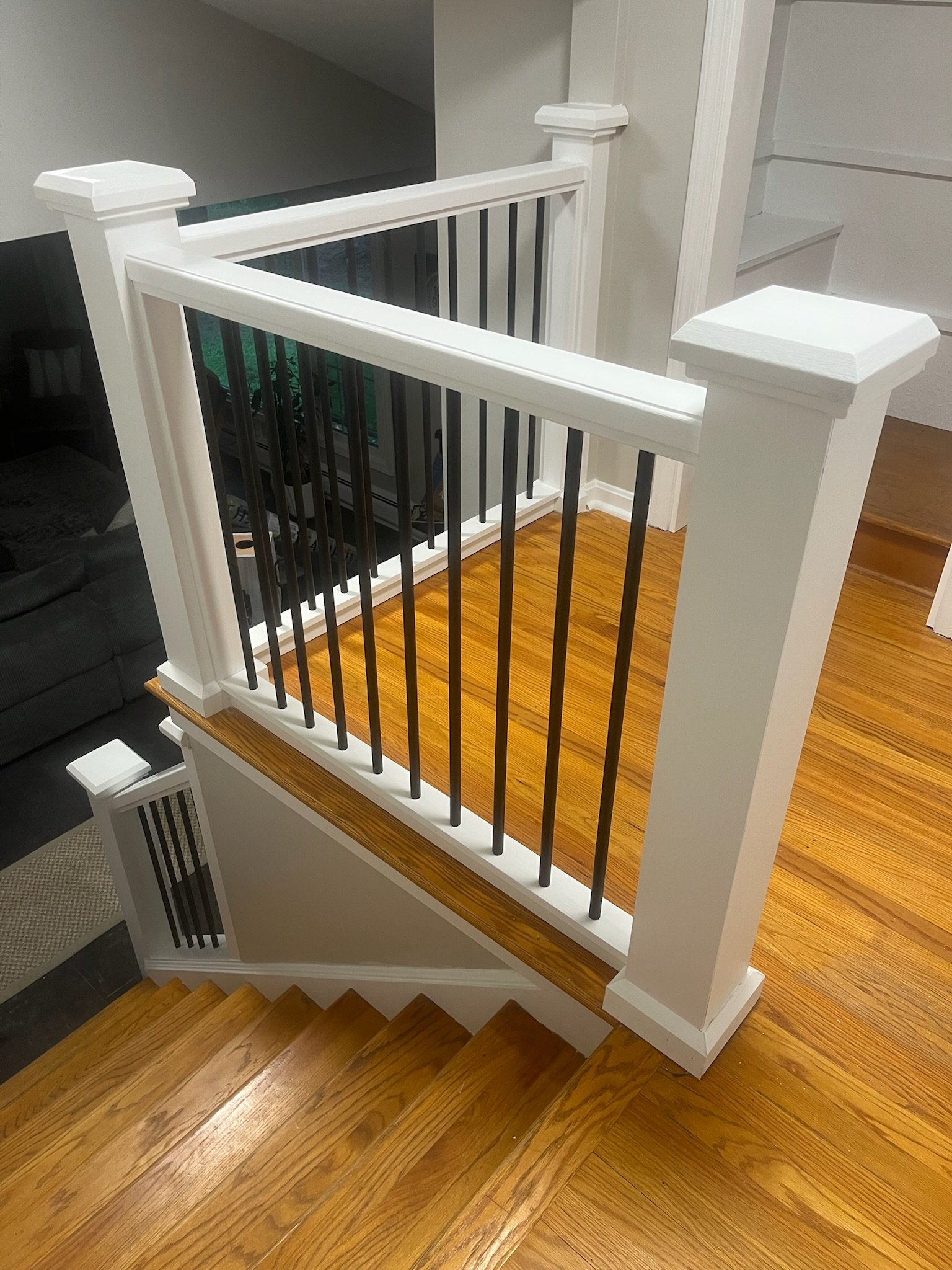 Interior view of a staircase with wooden steps, white painted railing and black metal balusters, leading down to a lower level with hardwood flooring.