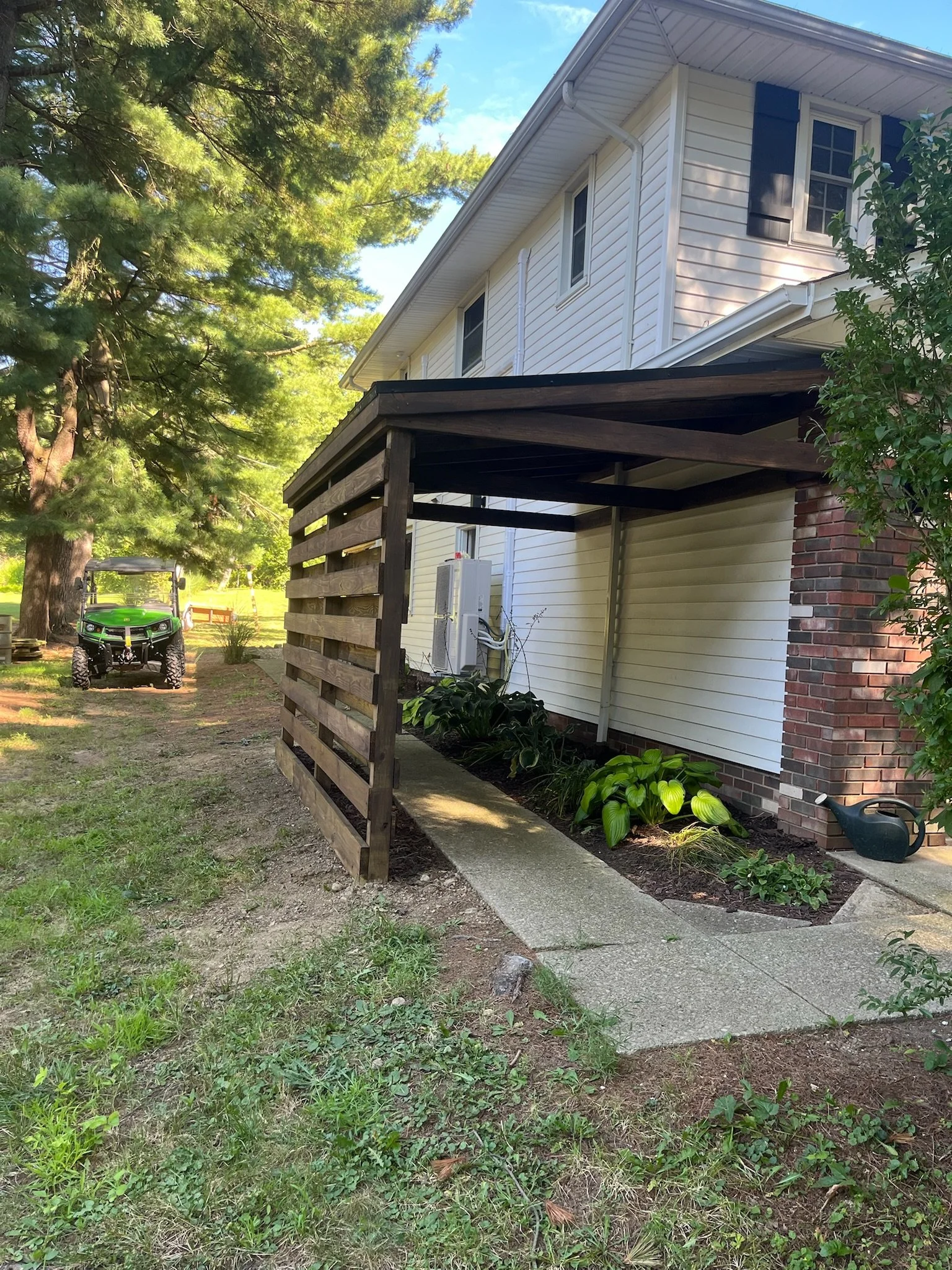 Side view of a house with a small porch, garden plants, a watering can, and a green utility vehicle in the yard, with a large tree nearby and bright blue sky. A new wooden awning has been built on the side.