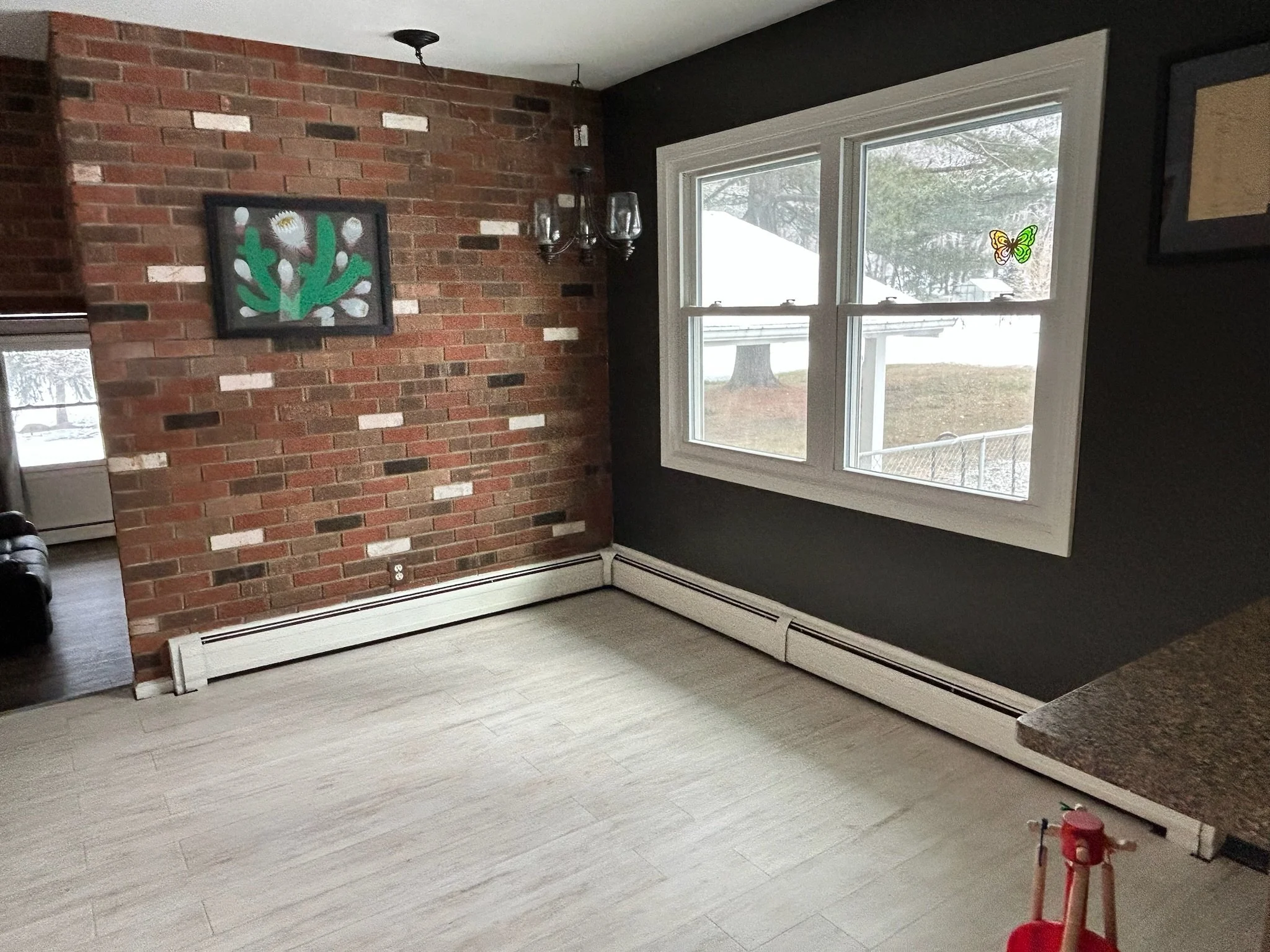 Empty room with brick accent wall, large window with two butterfly stickers, dark wall paint, and baseboard heater.