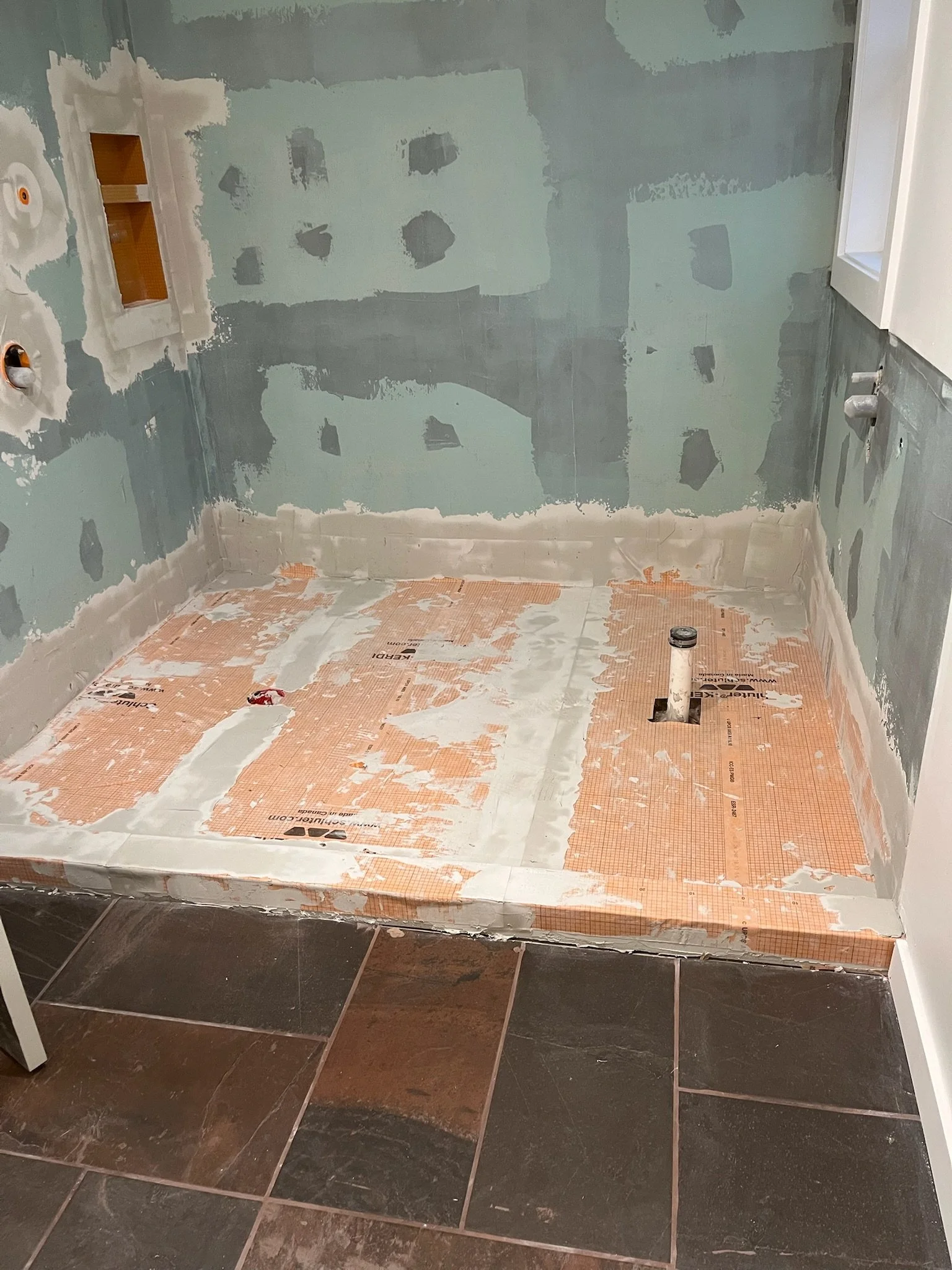 Bathroom under renovation showing unfinished drywall, plumbing pipes, and partial floor waterproofing installation.