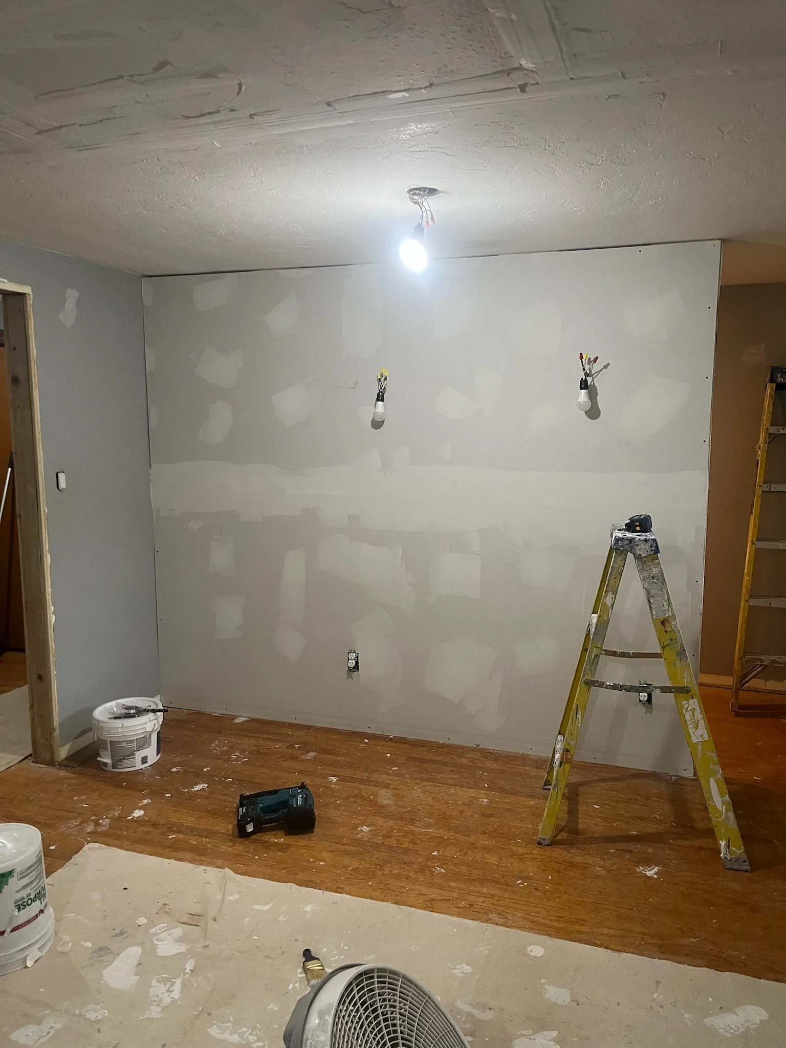 Room under renovation with drywall, ladder, construction tools, and a bright ceiling light.