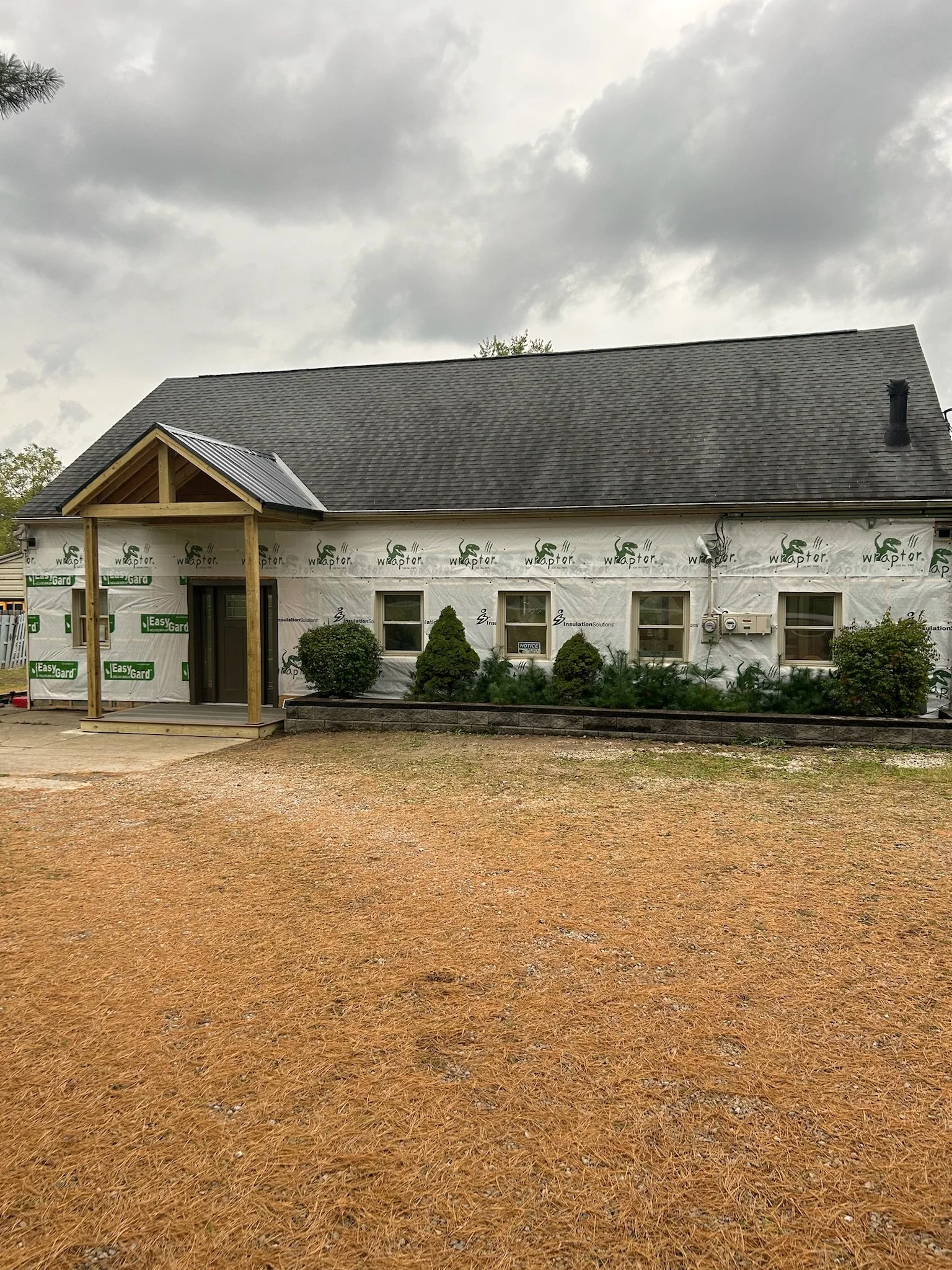 A house under construction with exterior walls wrapped in building material, a dark shingled roof, a front porch with new wooden supports, and landscaping with bushes and plants in front. The sky is cloudy.