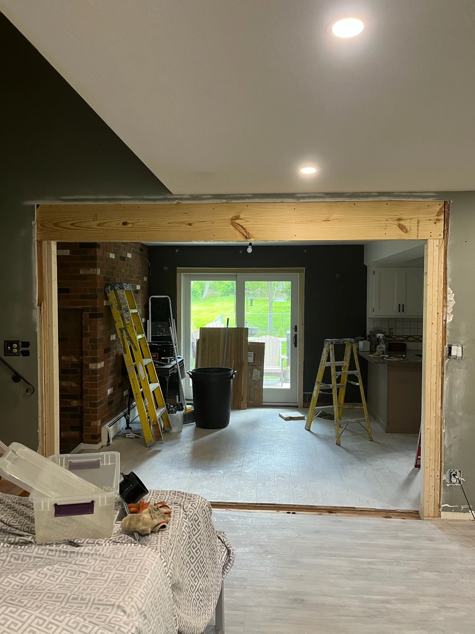 Living room undergoing renovation with new wooden framing around an opening, construction tools and ladders, a brick fireplace on the left, and a sliding glass door leading outside to a green yard.