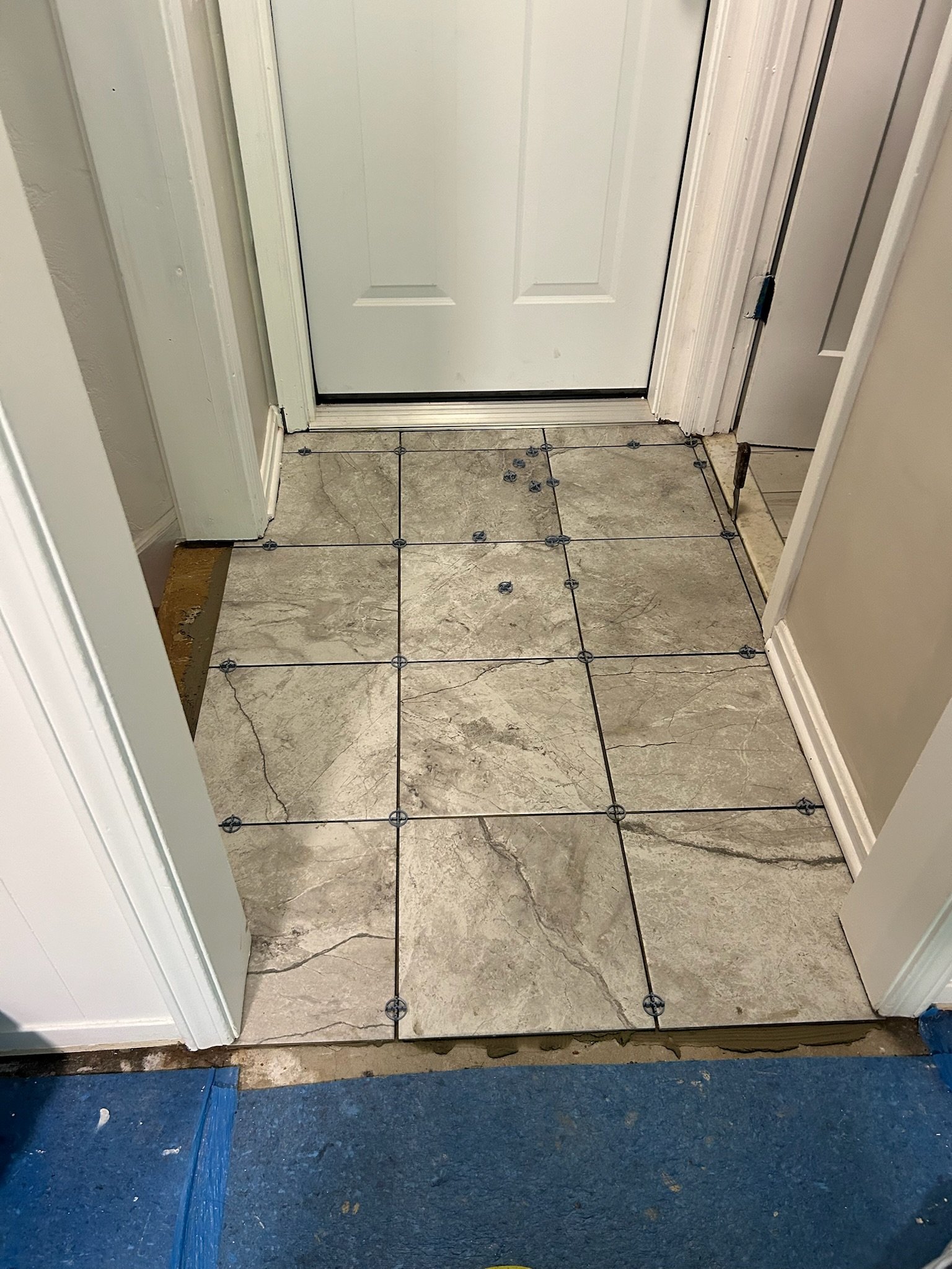 Inside view of a doorway with floor tiles being installed, showing tiles with gray marble pattern and spacing spacers in place.