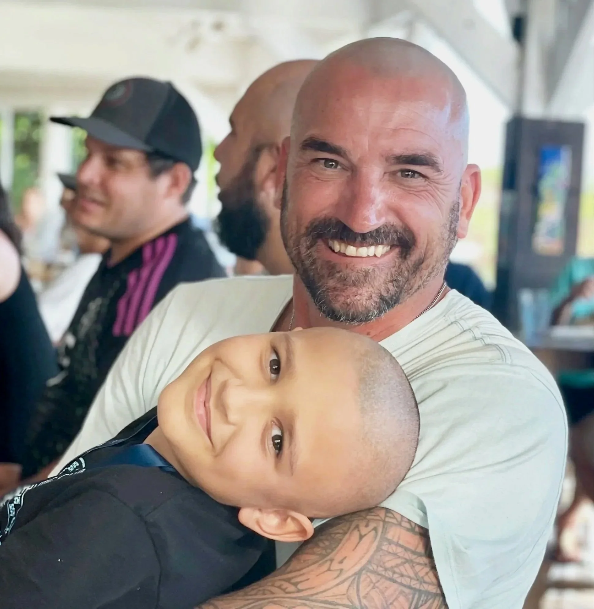 A man with a shaved head and beard smiling and holding a young boy with a shaved head, both showing a loving embrace in a cheerful setting.