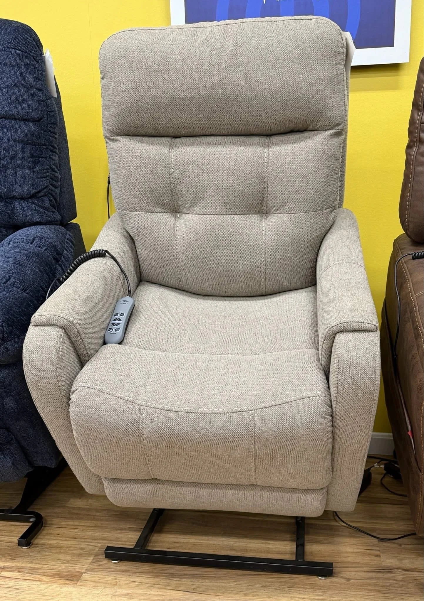 Beige fabric power lift recliner with a tall cushioned back, streamlined padded arms, stitched seat, and handheld remote.