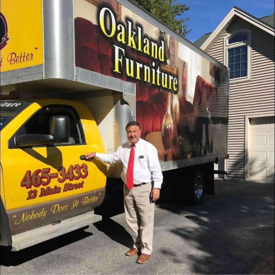 Representative of Oakland Furniture standing beside a branded delivery truck, highlighting family ownership and local roots in Oakland, Maine.