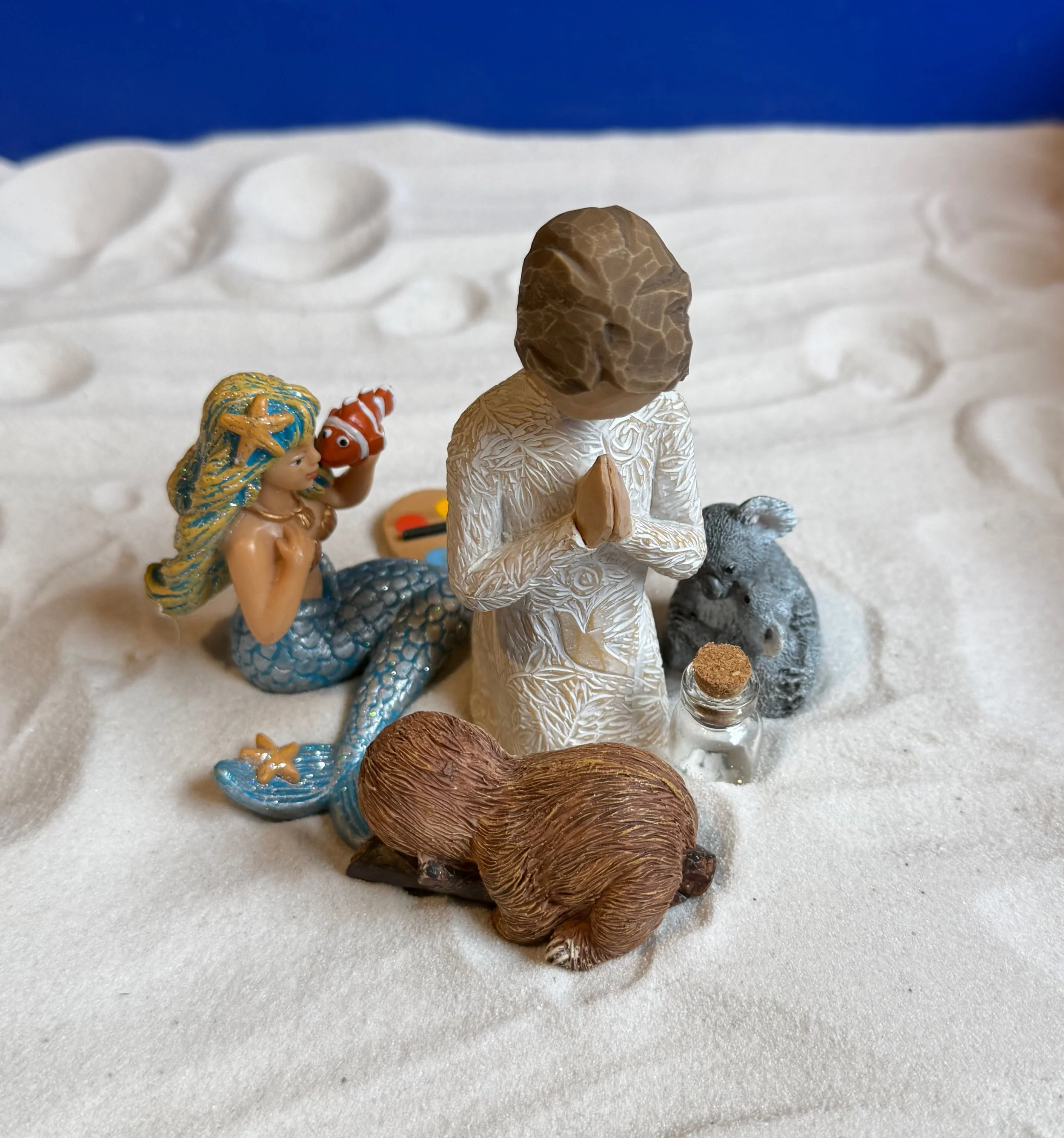 A nativity scene with a figurine of a praying angel, a mermaid, a small gray rabbit, a brown hamster, and a small bottle with a cork on a white textured surface.
