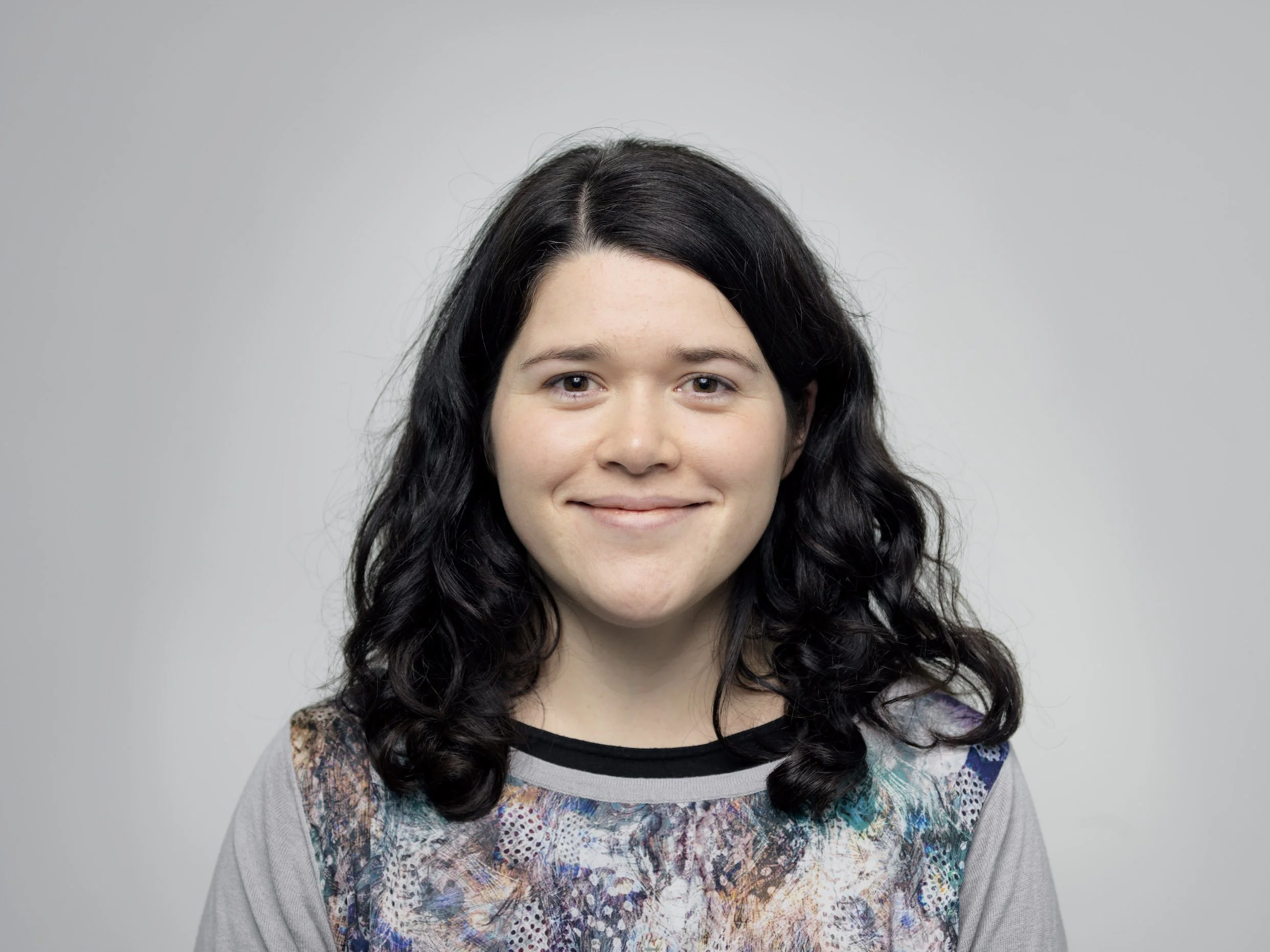 A young woman with black, wavy hair smiling against a plain gray background.